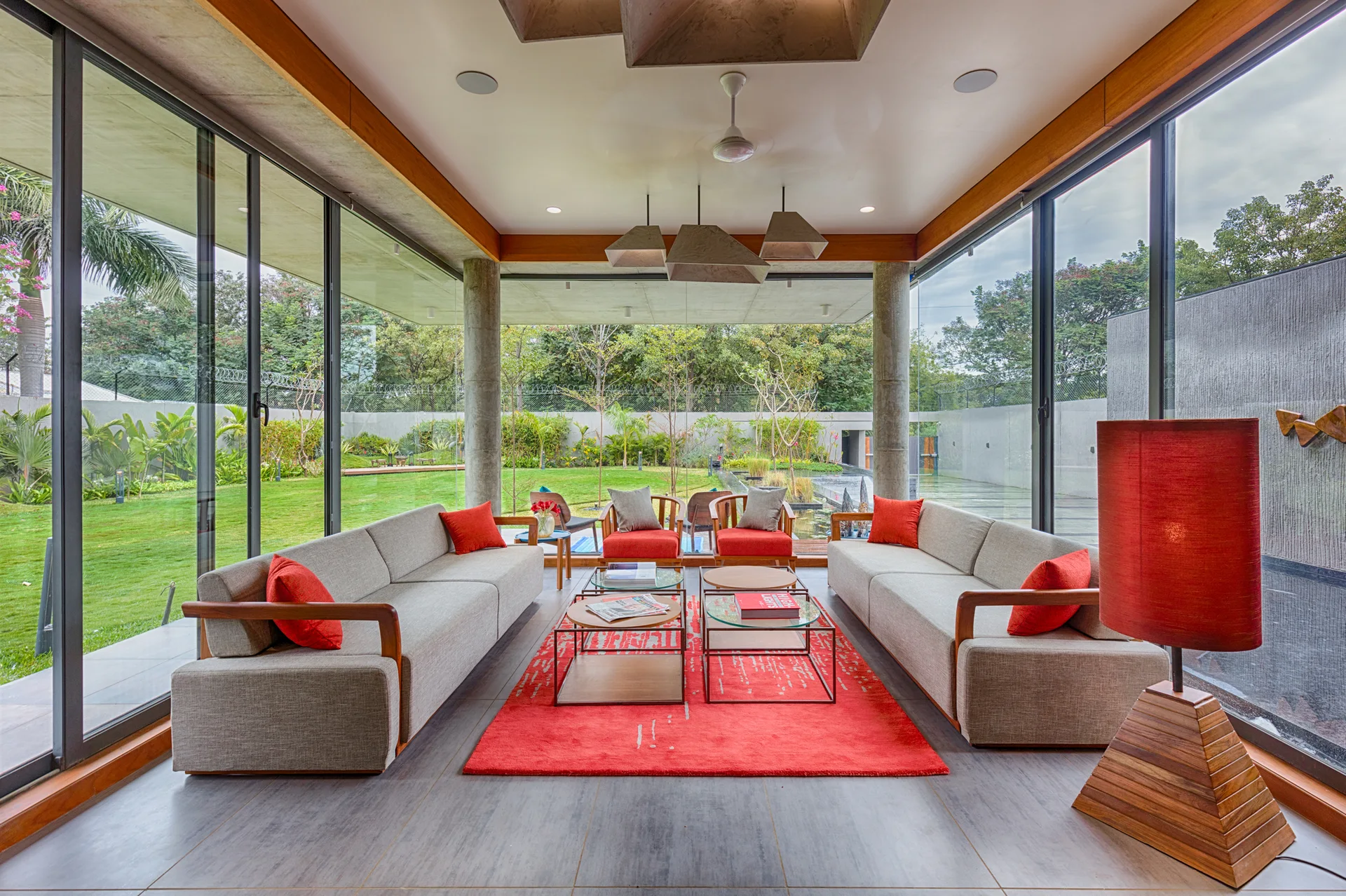 Two light grey sofas face each other on a large red rug inside a modern living space wrapped in glass walls.