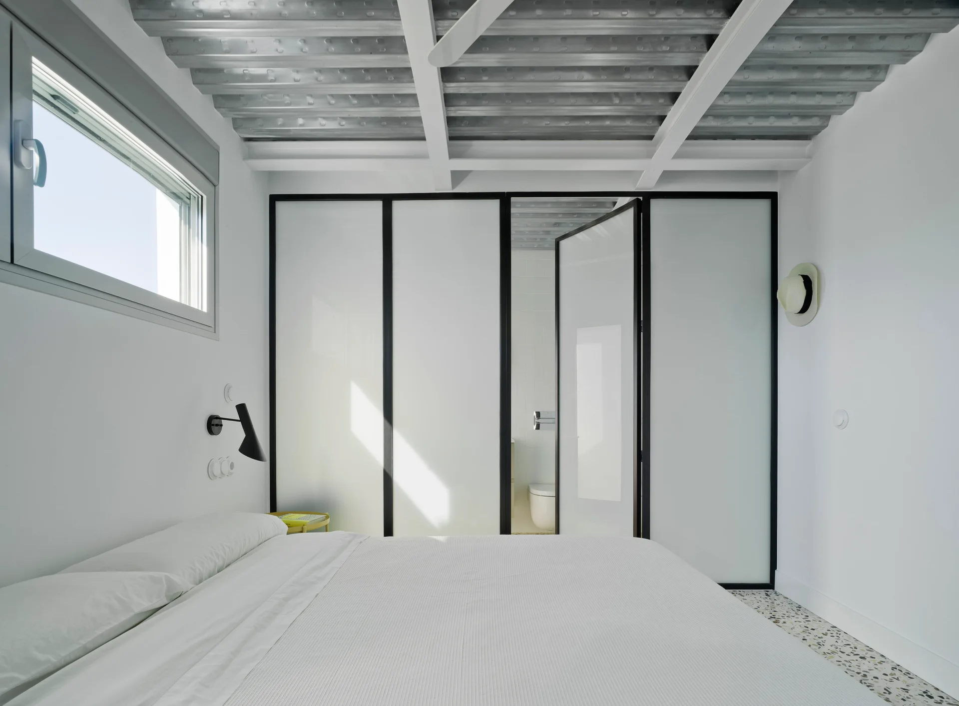A minimalist bedroom featuring an exposed corrugated metal ceiling, white bedding, terrazzo floors, and tall black-framed frosted glass partition doors.