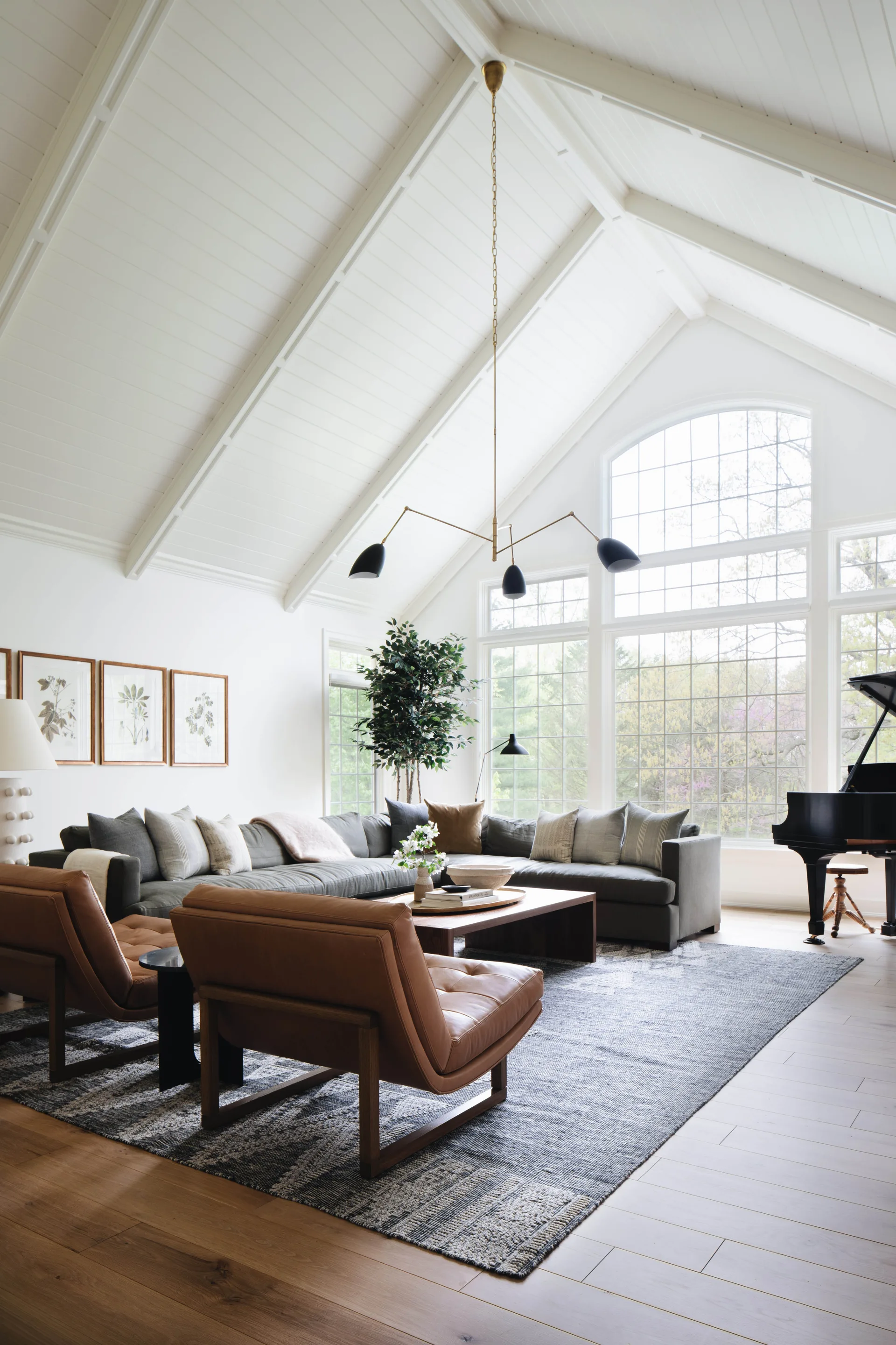 A spacious living room featuring a grey sectional, two tan leather armchairs, and a black grand piano under a high white vaulted ceiling.