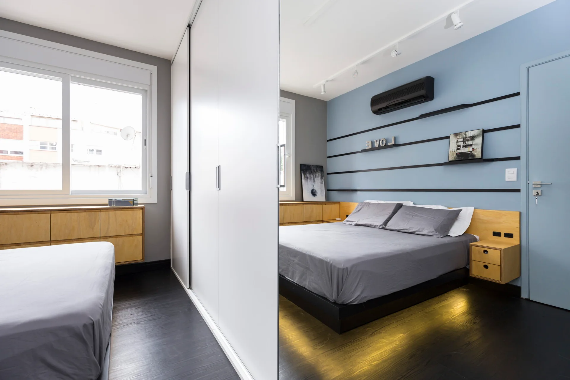 A modern bedroom features a platform bed with grey linens, an integrated wood headboard, a blue accent wall, and white sliding wardrobe doors.