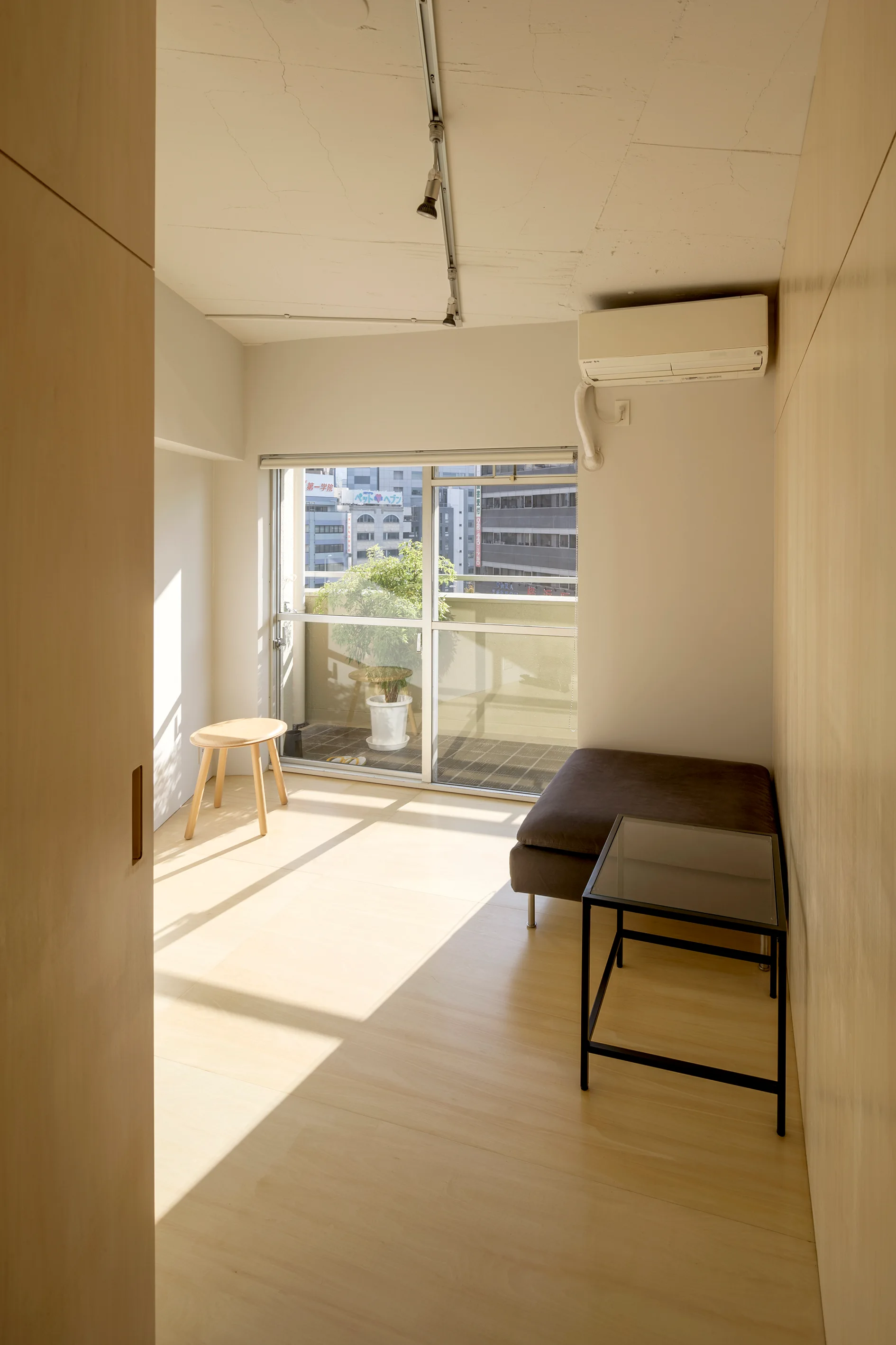 A small minimalist room with light wood wall paneling, matching floors, a low grey lounge cushion, and a glass-top table next to a sliding balcony door.