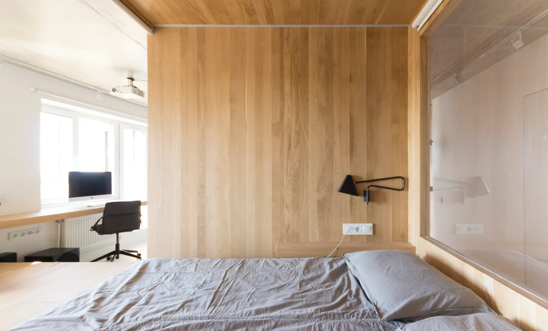 A minimalist bedroom features vertical light wood paneling, a grey bed, and a glass partition wall looking into a bright home office.