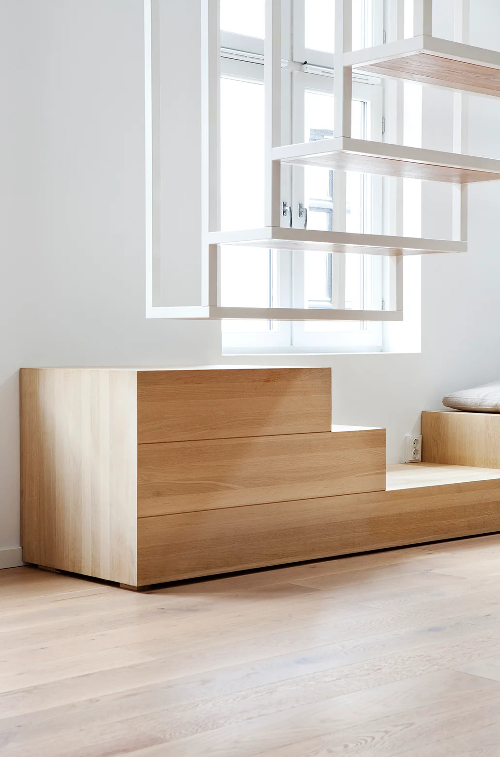 A custom stepped light wood cabinet sits on matching flooring below a white open-frame hanging staircase next to a window.