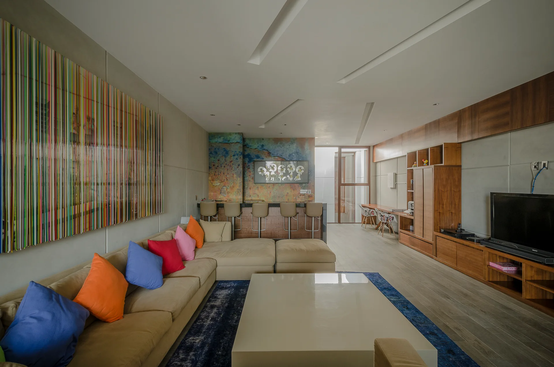 A modern living room features a tan sectional sofa with colorful pillows, a white coffee table on a blue rug, and built-in wood cabinetry.