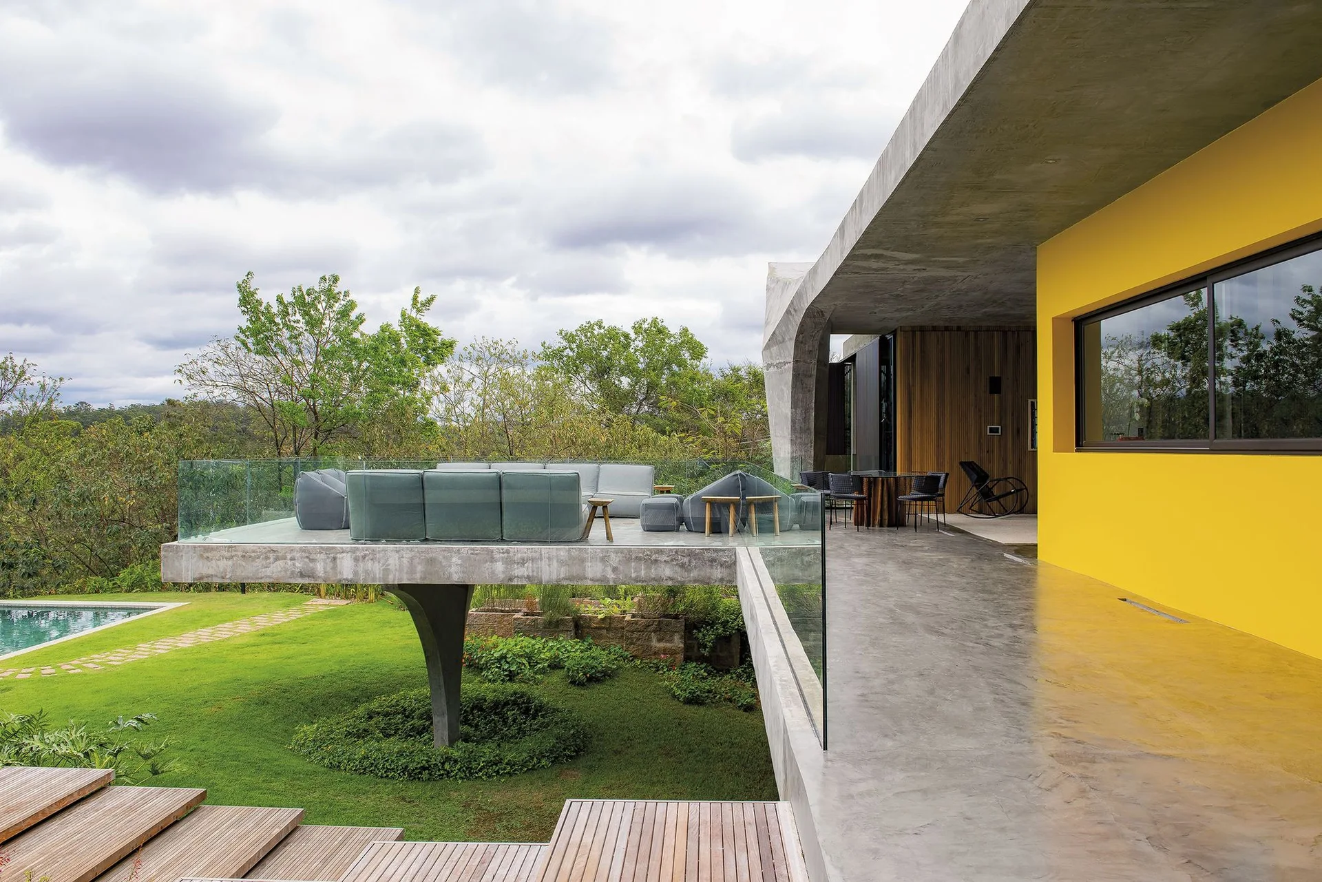 A cantilevered concrete terrace features a grey modular sectional sofa, a glass railing, a bright yellow exterior wall, and wooden steps.