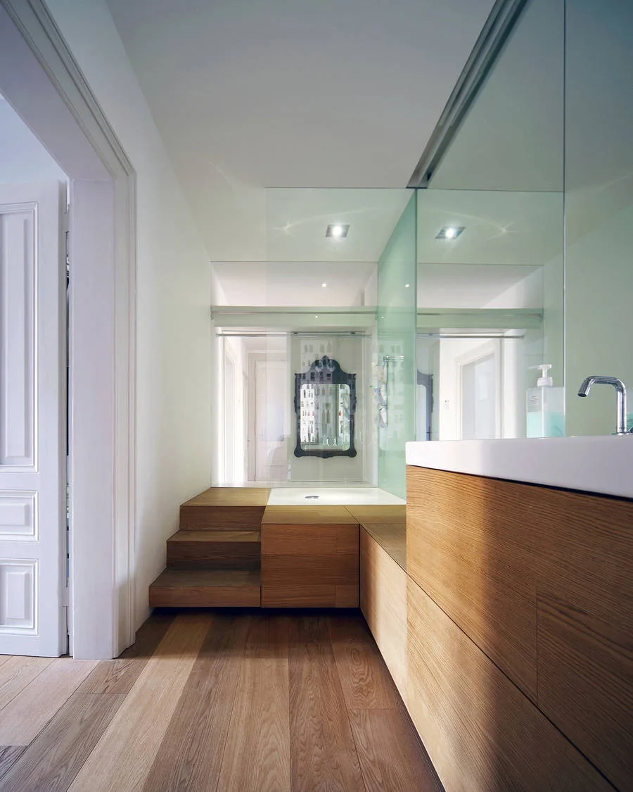 A minimalist bathroom featuring a light wood vanity and matching steps that lead up to a raised platform behind a glass screen.
