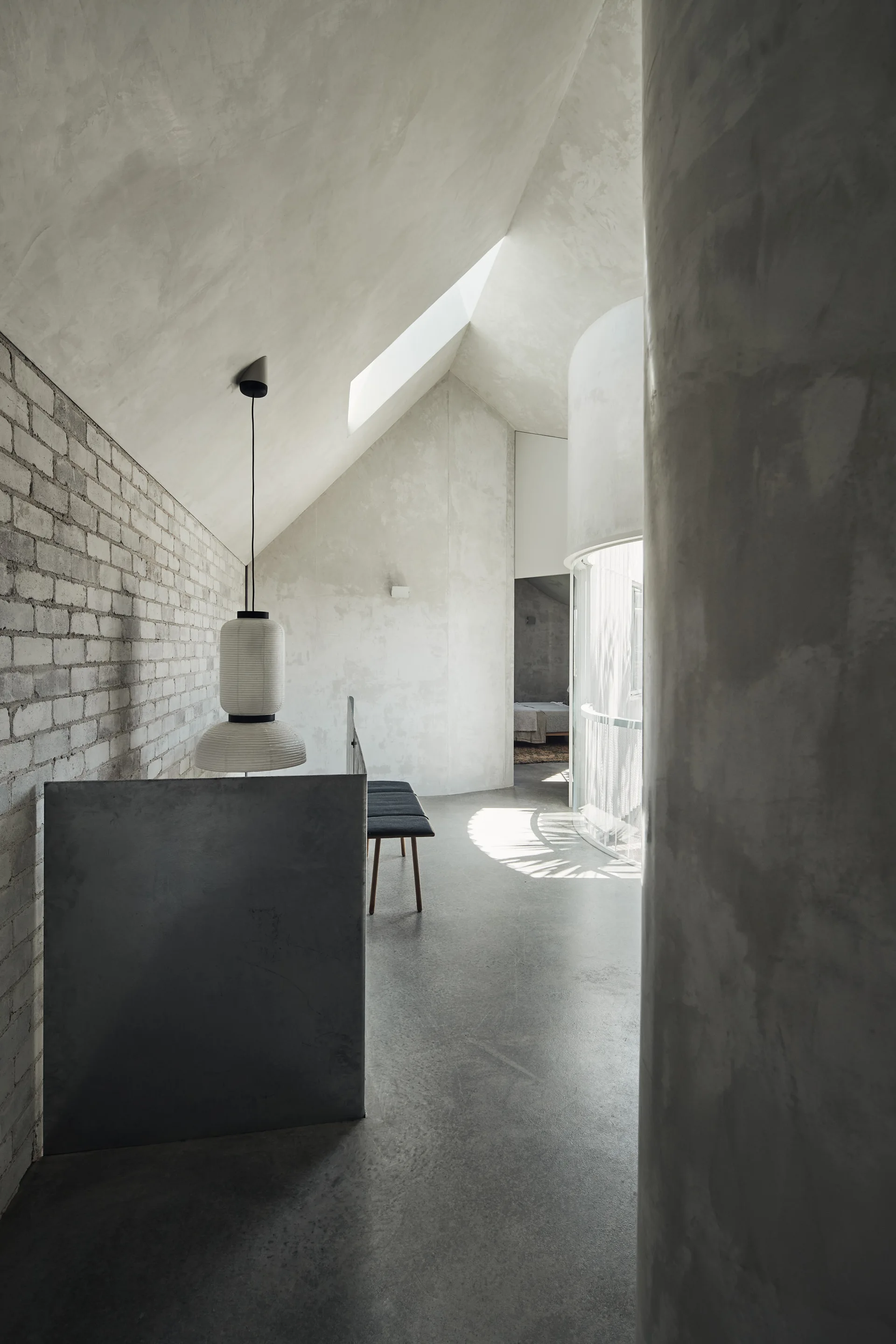 A minimalist grey concrete landing features an exposed brick wall, a sloped ceiling with a skylight, and a hanging paper lantern pendant light.