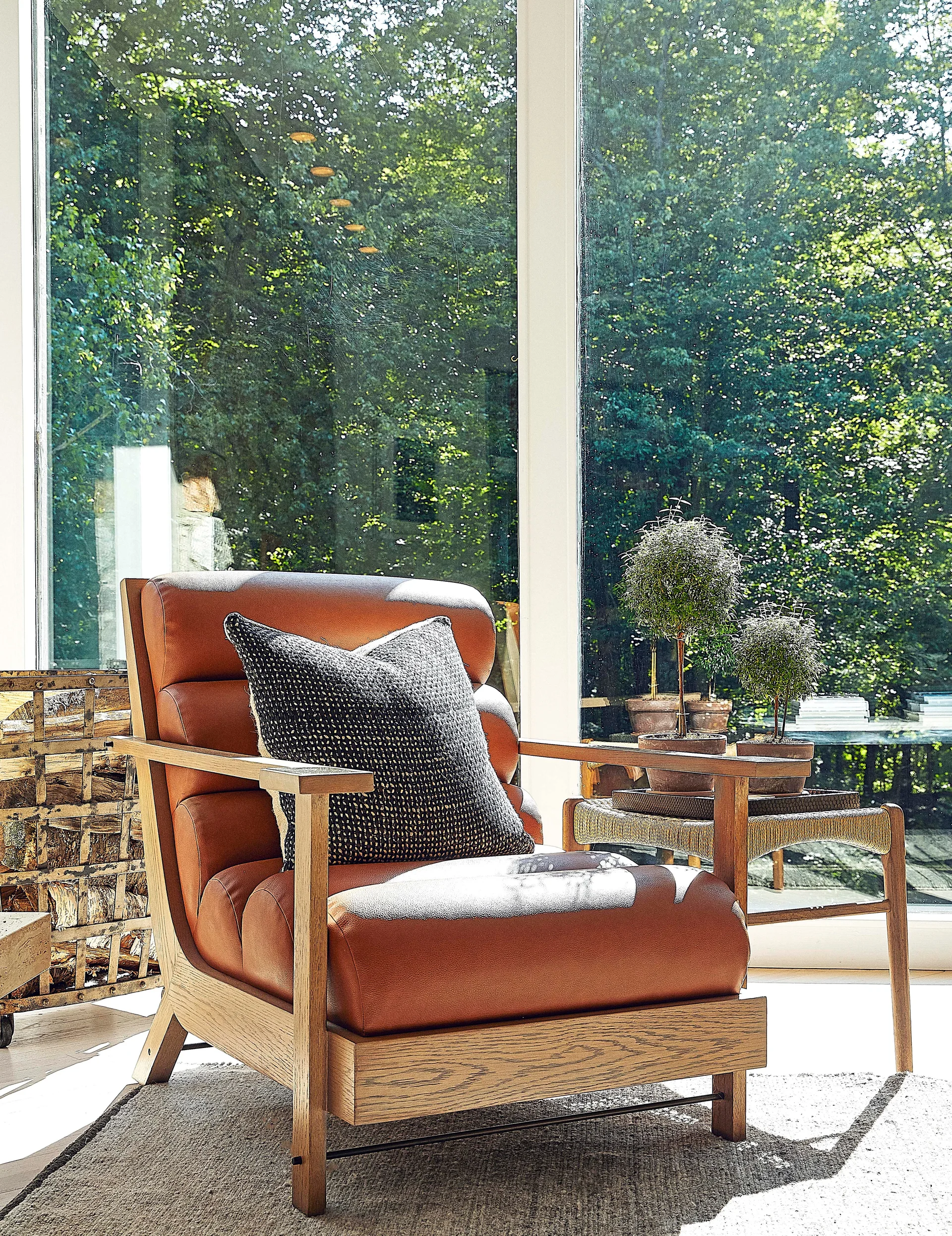 A brown leather armchair with a dark pillow sits by a woven wooden side table in front of a large window overlooking trees.