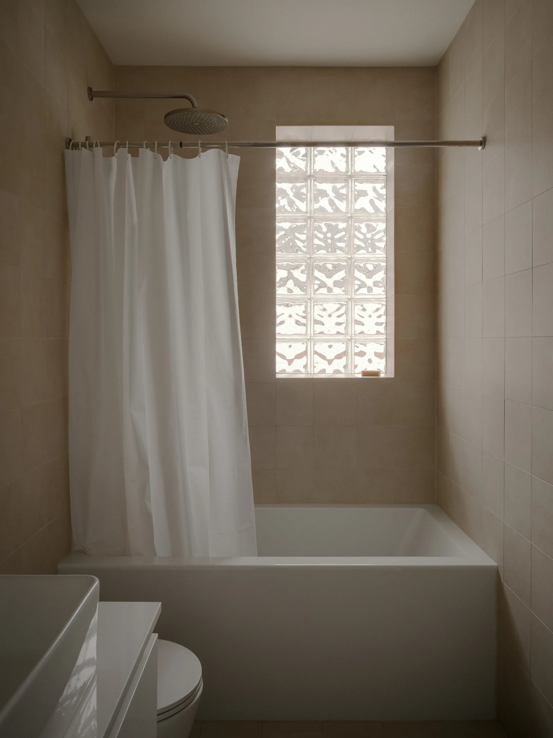 A beige tiled bathroom features a white bathtub, a matching white shower curtain, and a vertical glass block window.