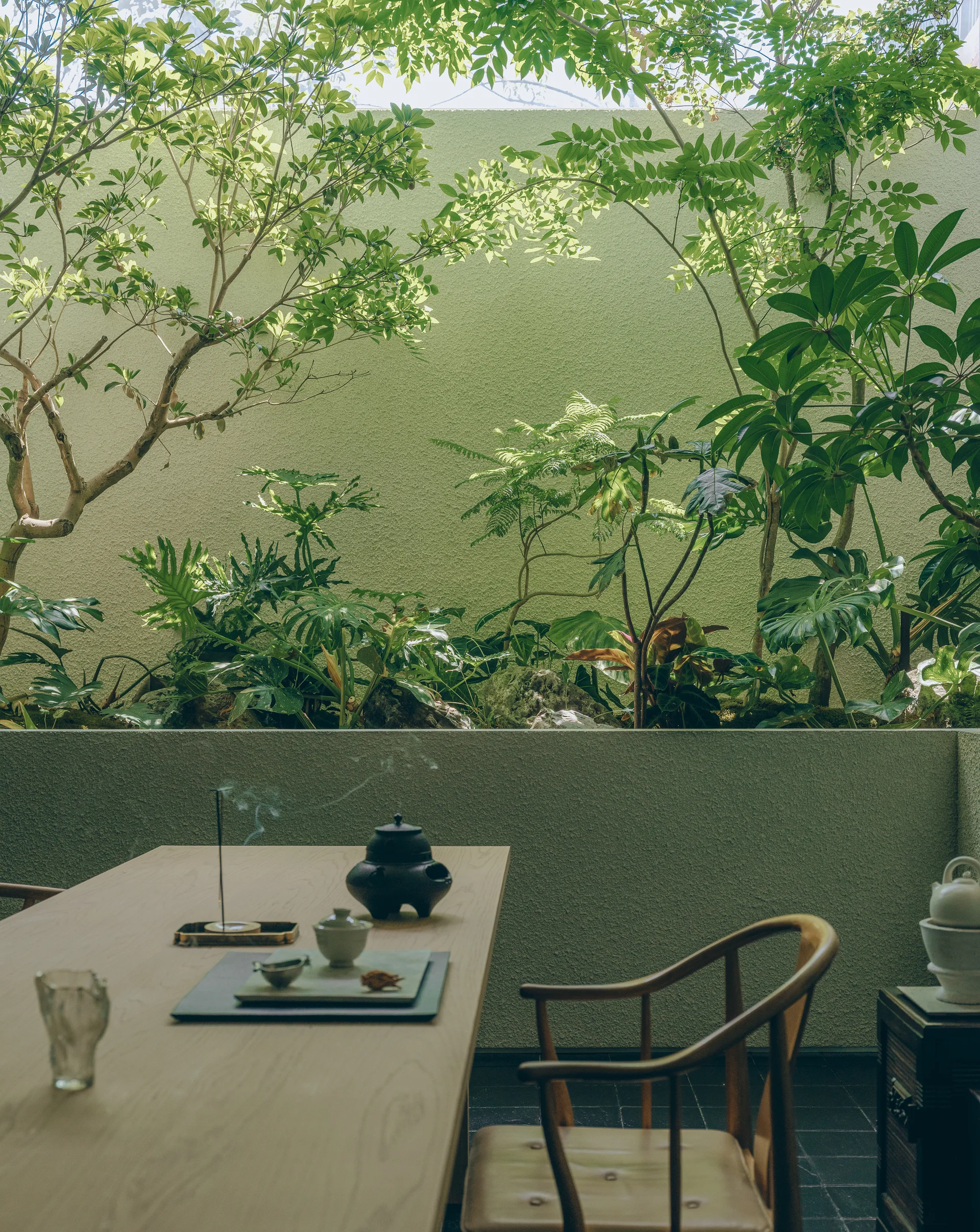 A light wood table set with a black teapot and ceramic cups sits in a tiled courtyard next to a green retaining wall filled with lush plants.