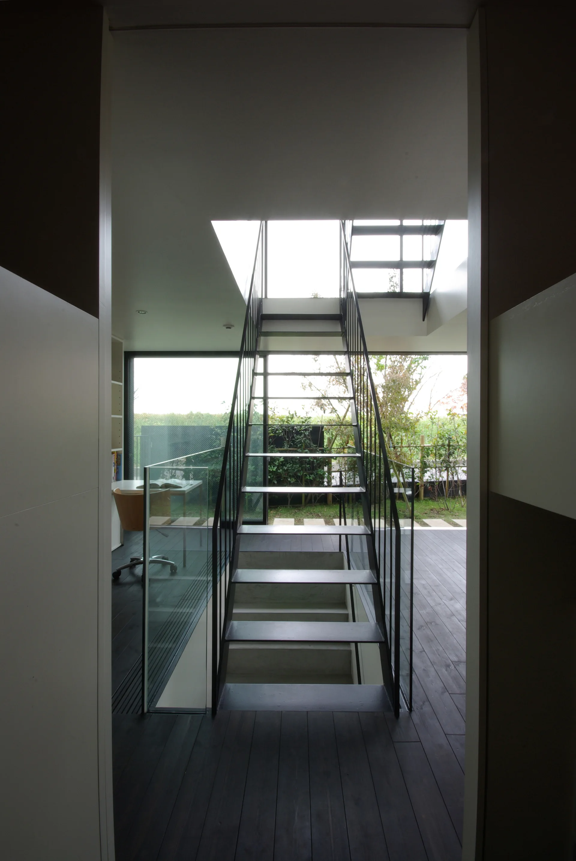 A minimalist home interior featuring a central black metal staircase with open treads, dark wood flooring, and a large window looking onto a garden.