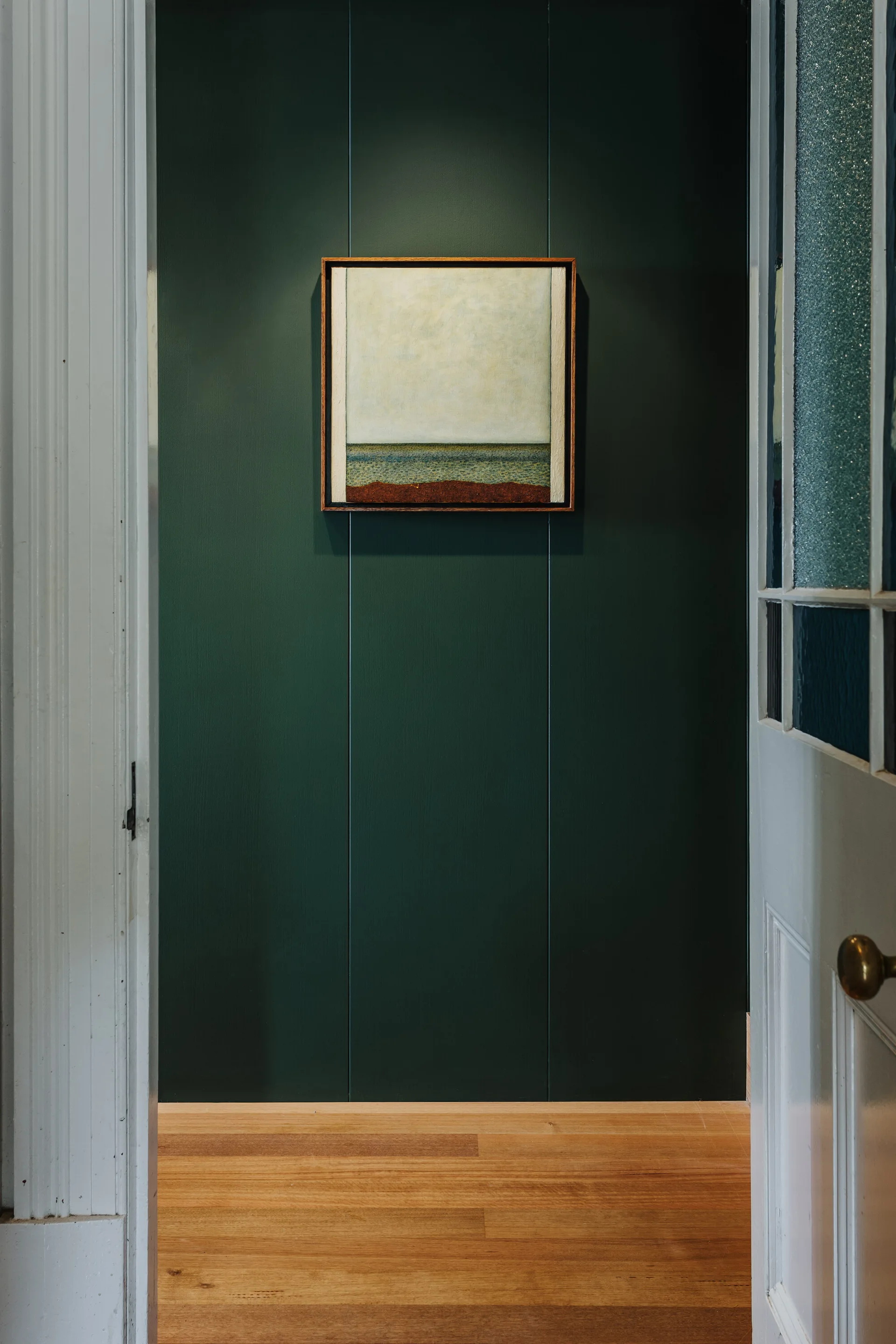 A view through a white doorway onto a dark green paneled wall featuring a framed square painting illuminated by a spotlight over hardwood floors.