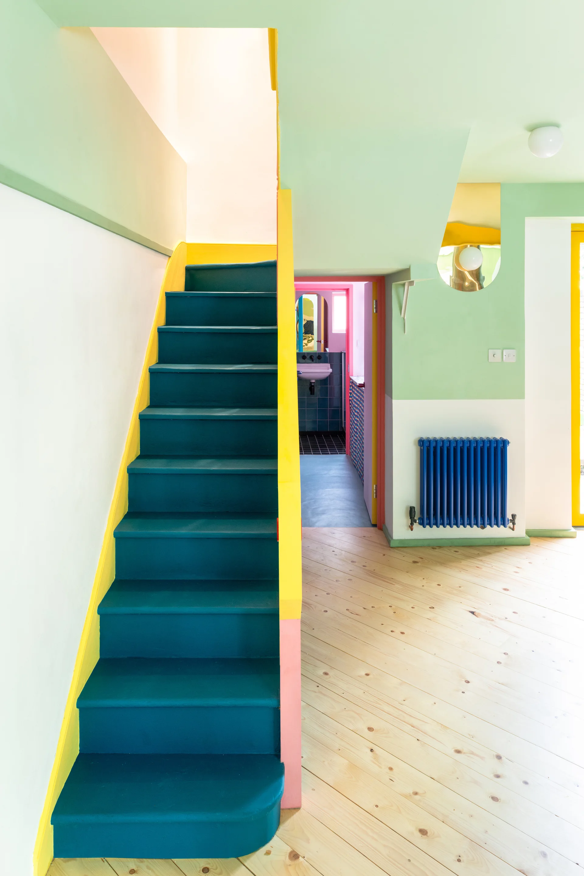 A teal painted staircase with yellow trim stands on light wood floors next to a blue radiator and color-blocked mint green and white walls.