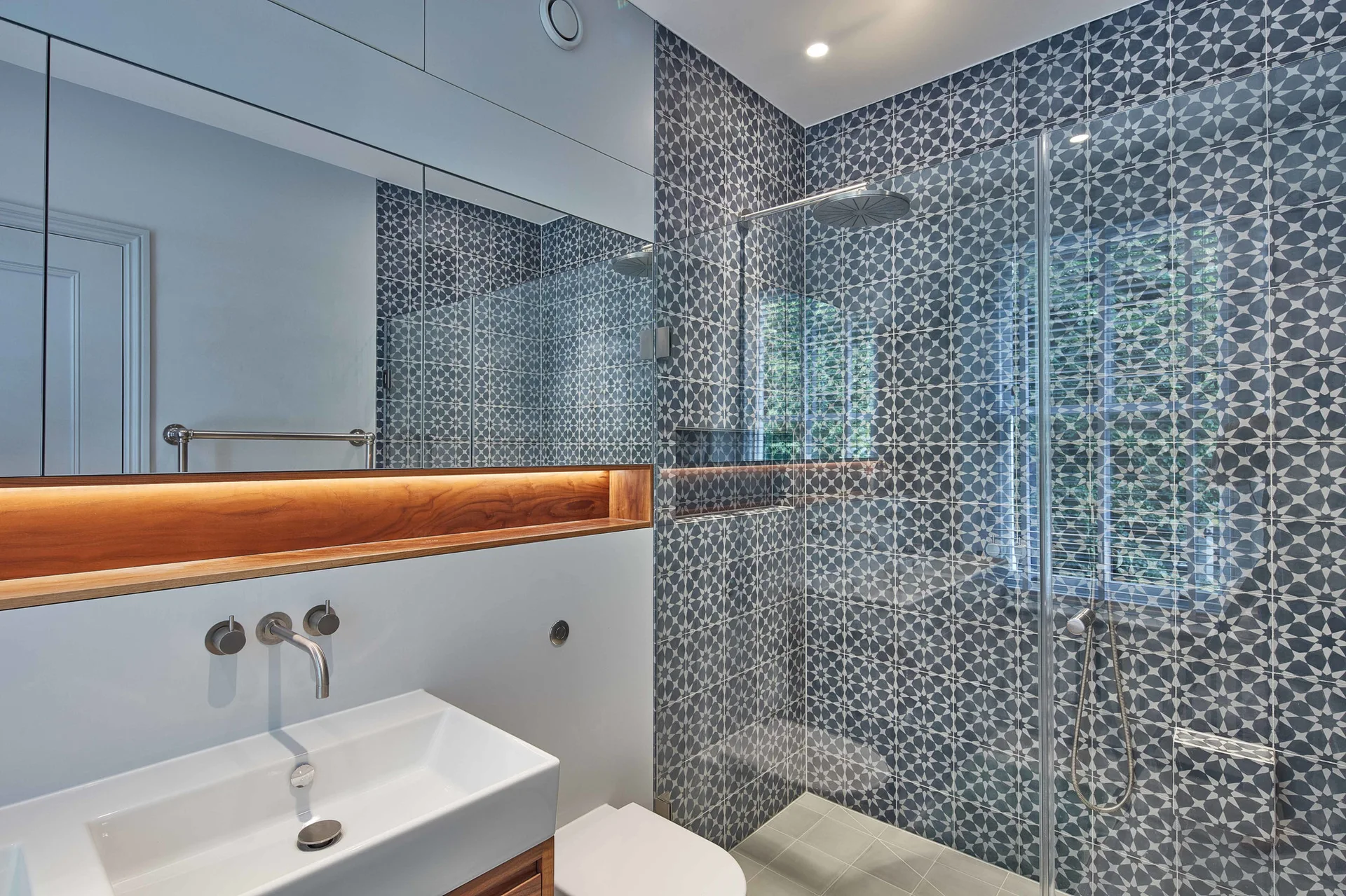 A modern bathroom features a glass-enclosed walk-in shower with geometric blue tiles next to a white sink and a lighted wall mirror.