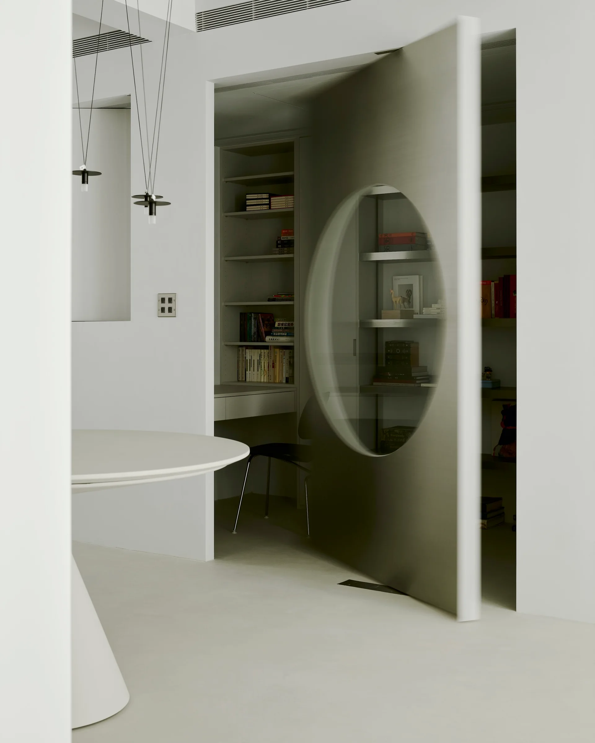 An open grey metal pivot door featuring a circular window leads into a white minimalist home library with built-in shelves and a desk.