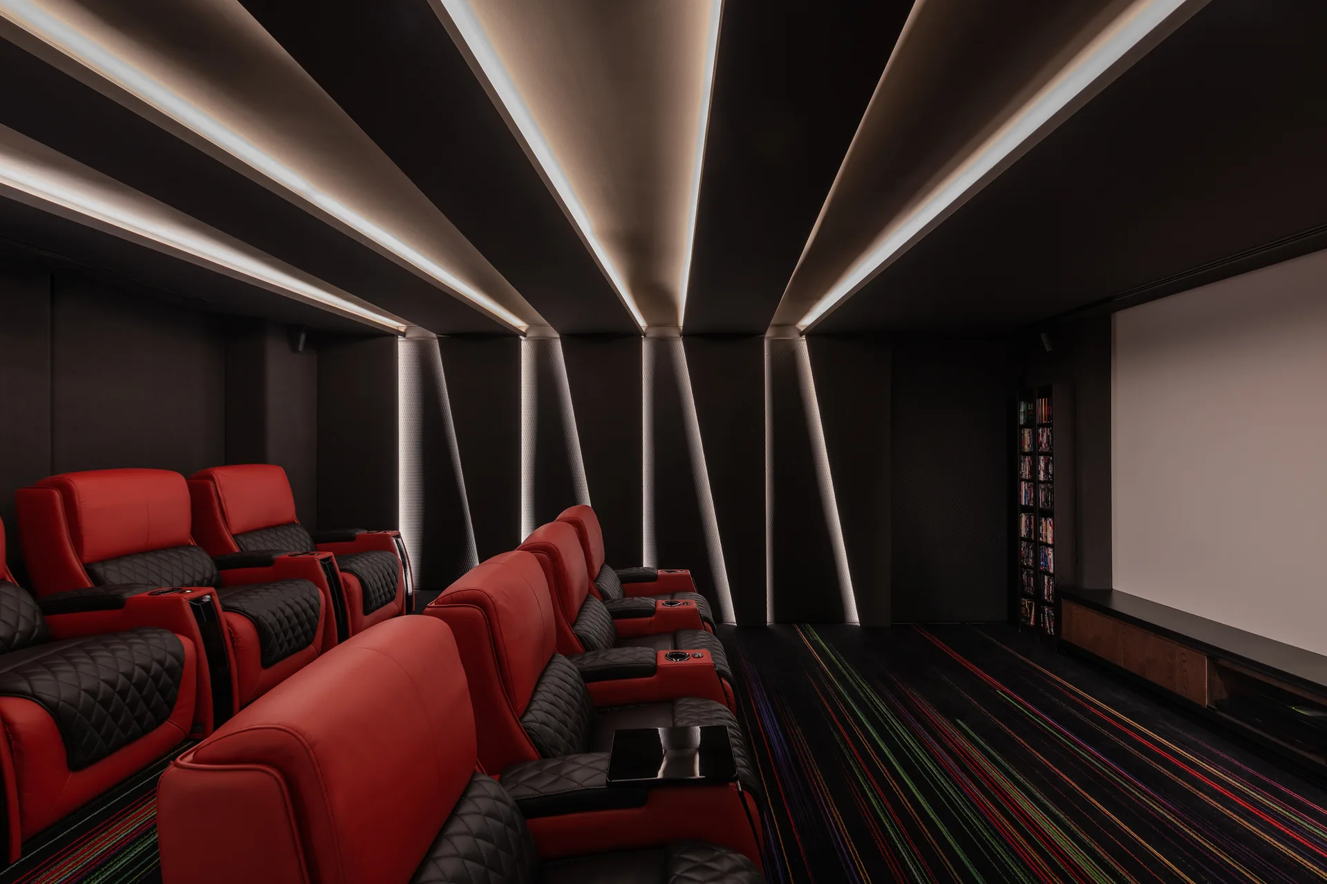 Red and black leather recliners are arranged in rows inside a dark home theater featuring a large screen and geometric, illuminated wall panels.