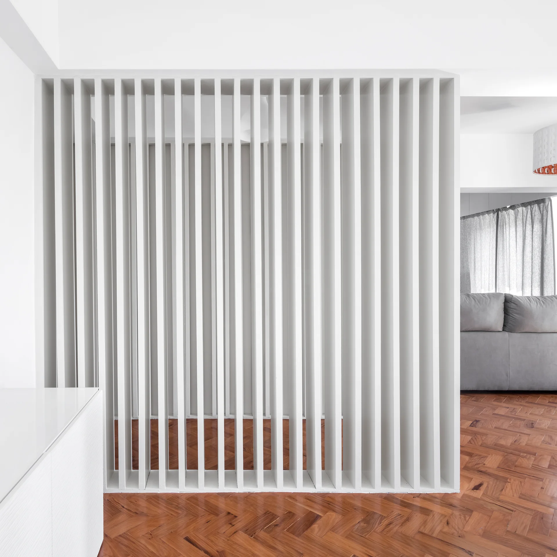 A vertical white slatted room partition stands on a rich herringbone wood floor, revealing a grey sofa and sheer curtains in the background.