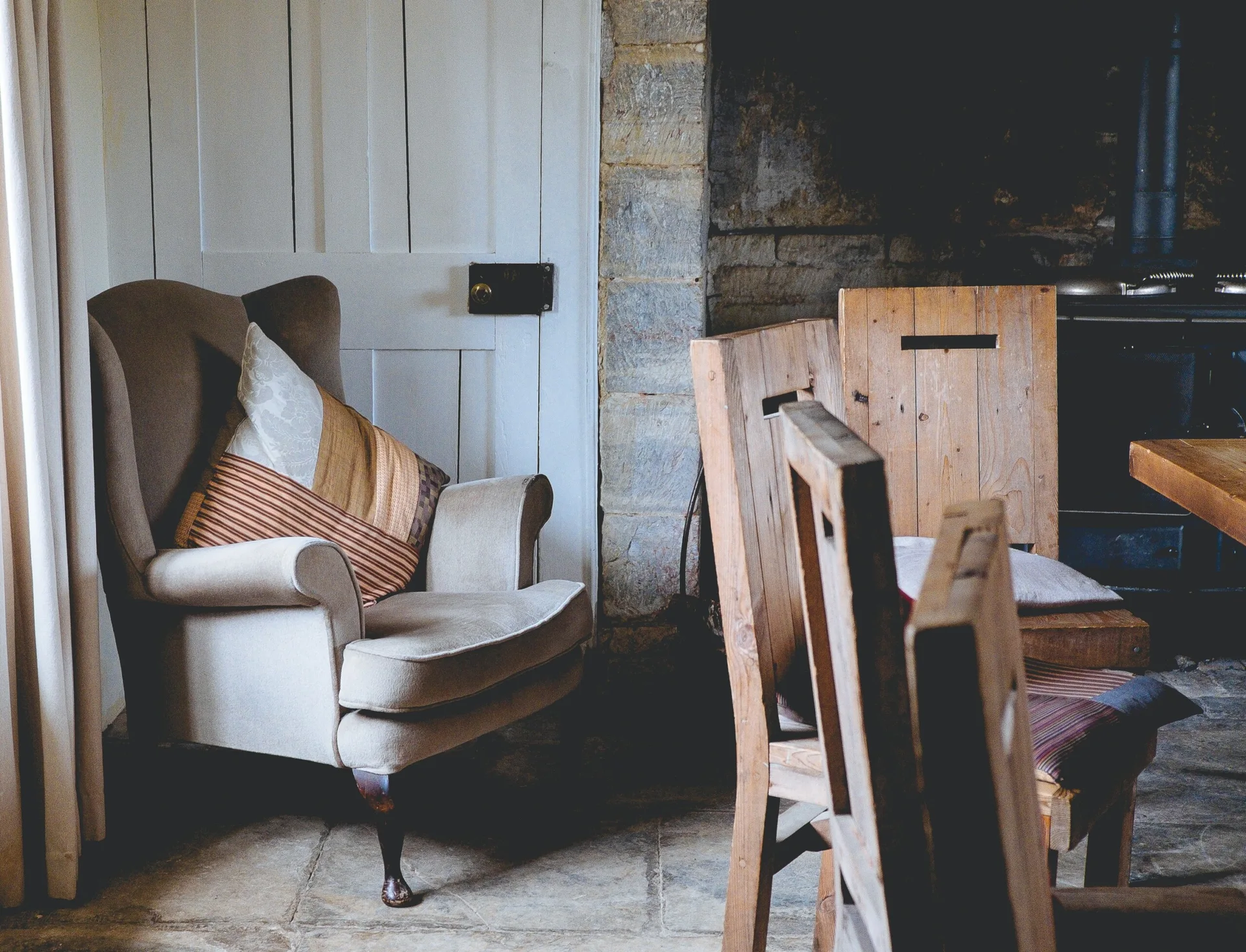 An upholstered beige wingback chair with a striped pillow sits next to a rustic stone wall, a white paneled door, and wooden chairs.