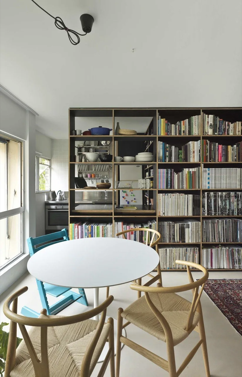 A bright dining room features a round white table, woven-seat wooden chairs, a blue high chair, and a large grid bookshelf acting as a room divider.