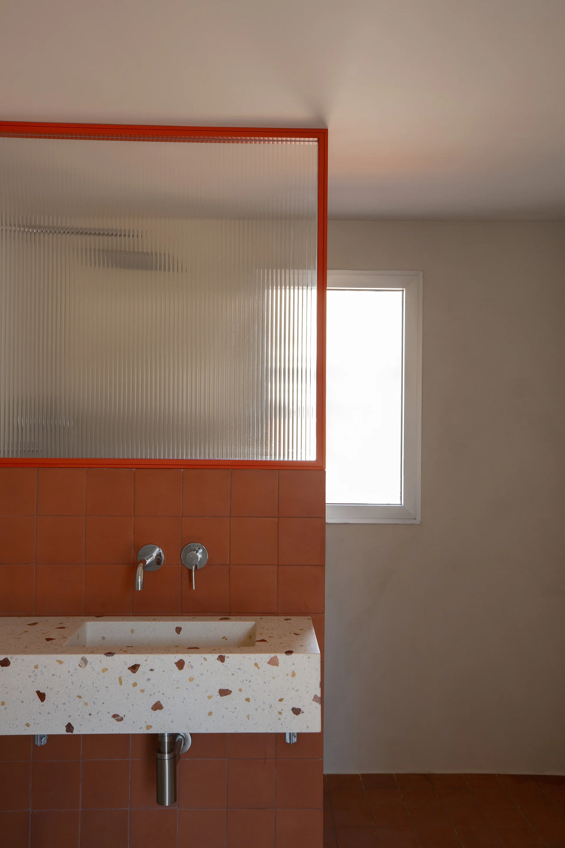 A modern bathroom features a speckled terrazzo sink mounted against terracotta wall tiles, below a ribbed glass screen with a bright orange frame.