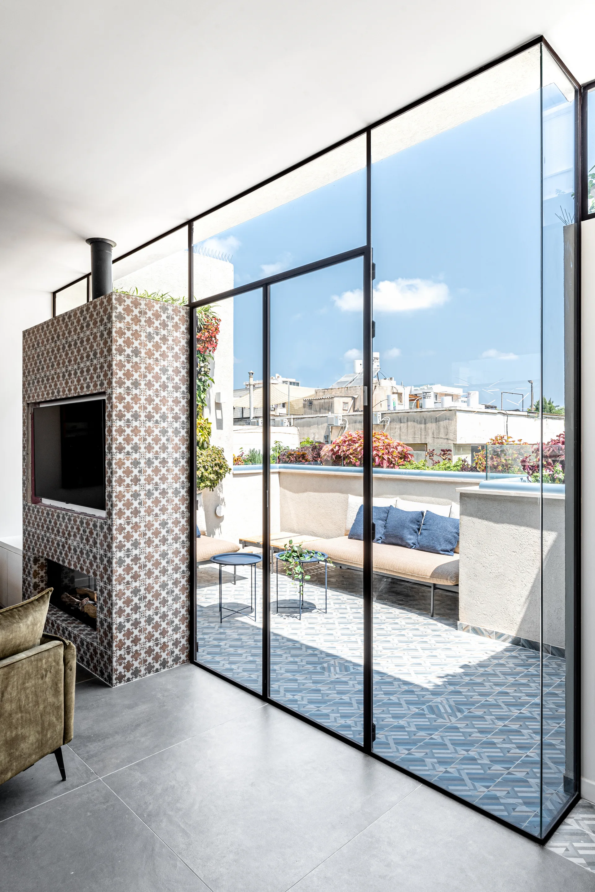 A modern living space features a patterned tile fireplace column and a large black-framed glass partition opening onto a sunny outdoor terrace.
