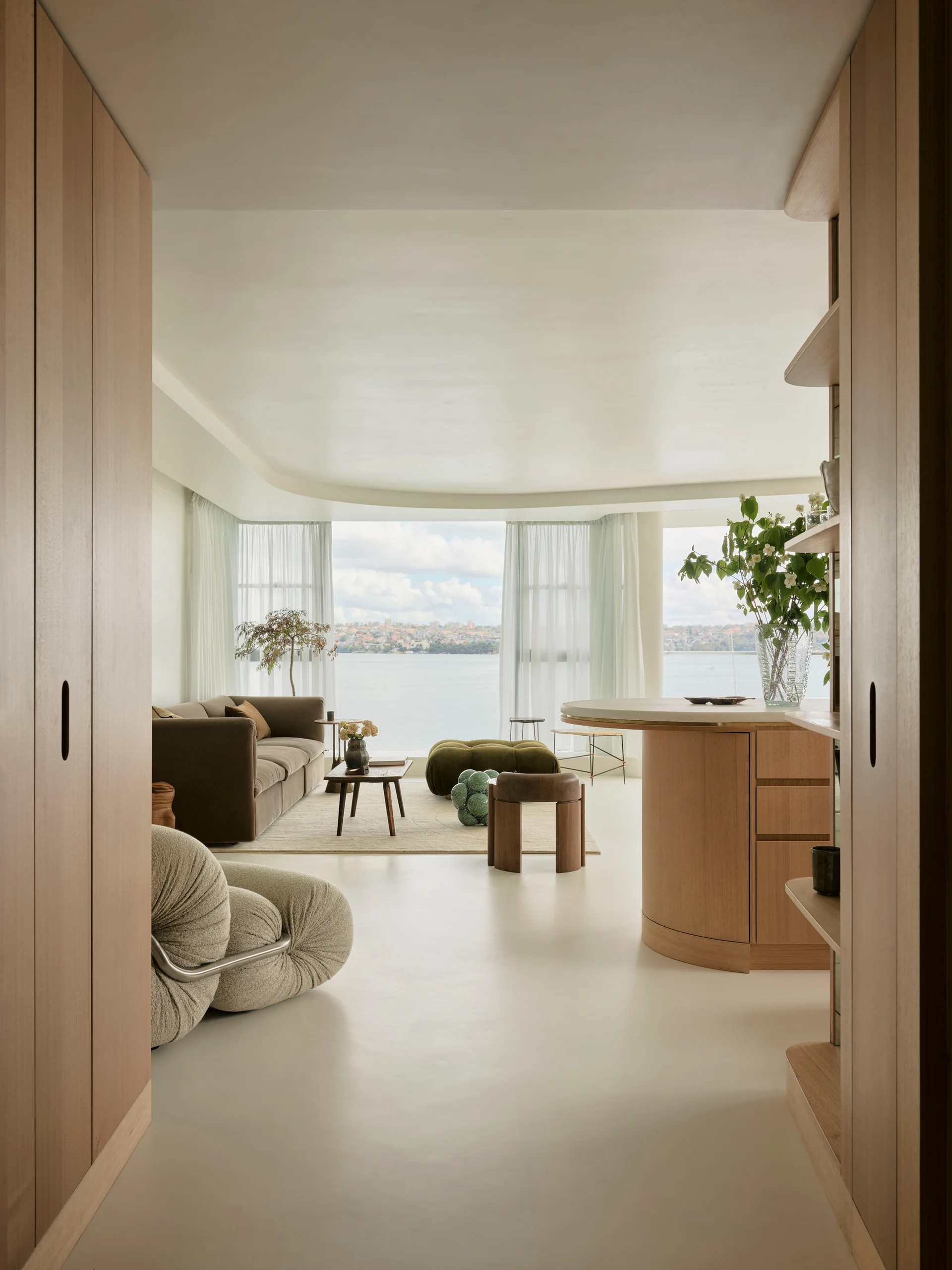 An open-plan living space with a curved wood island, brown sofa, and sheer curtains framing a panoramic waterfront view.