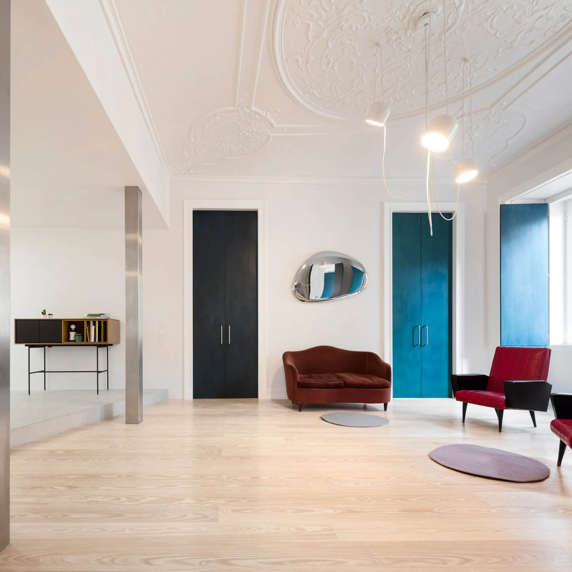 A spacious white living room with light wood floors, an ornate ceiling medallion, a burgundy sofa, and tall black and teal doors.