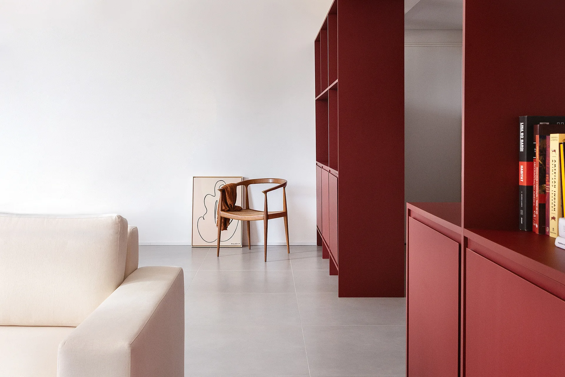A minimalist living space with a cream sofa, a wooden rattan armchair, and a large matte maroon shelving unit against a white wall.