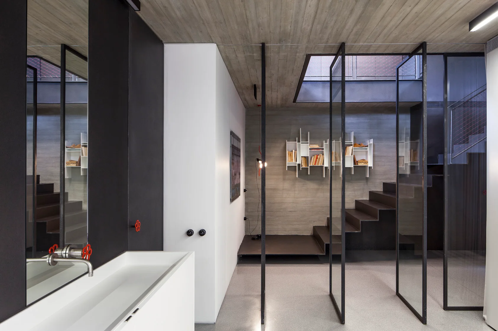 An industrial minimalist interior with a raw concrete ceiling, a dark metal staircase, glass partition panels, and a white sink.