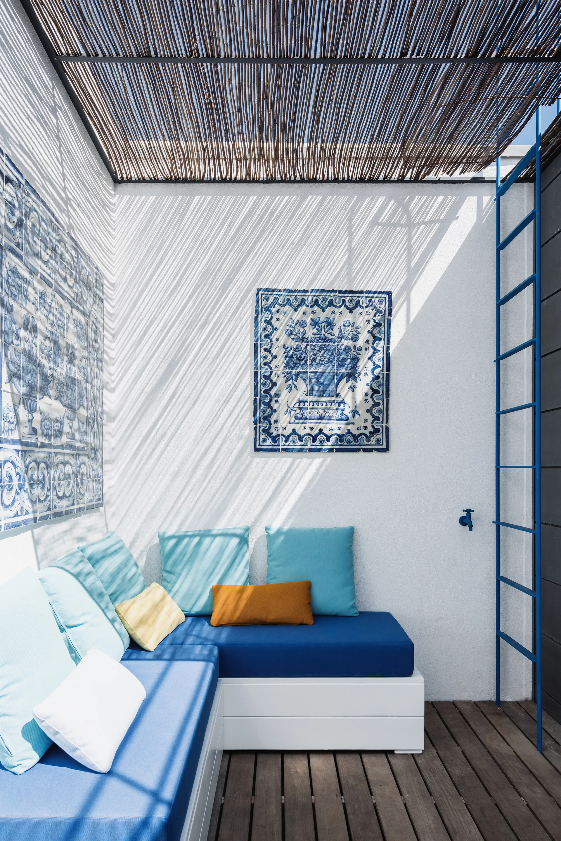 An outdoor patio features a white built-in corner bench with blue cushions under a slatted reed ceiling next to blue and white wall tiles.