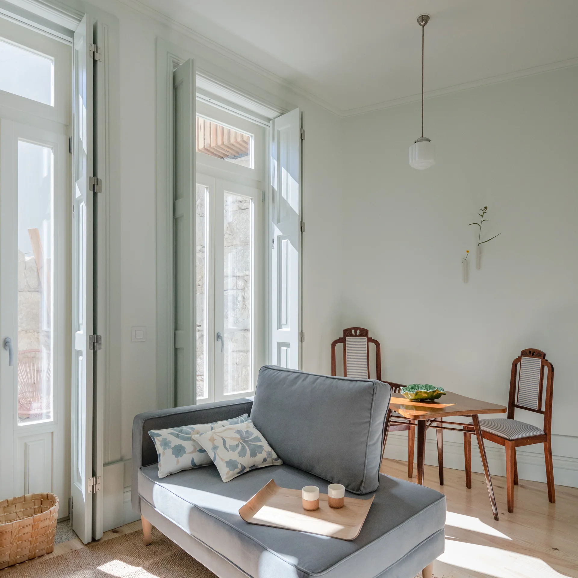 A sunlit room features a grey upholstered sofa, pale green interior window shutters, and a wooden dining table with two chairs.