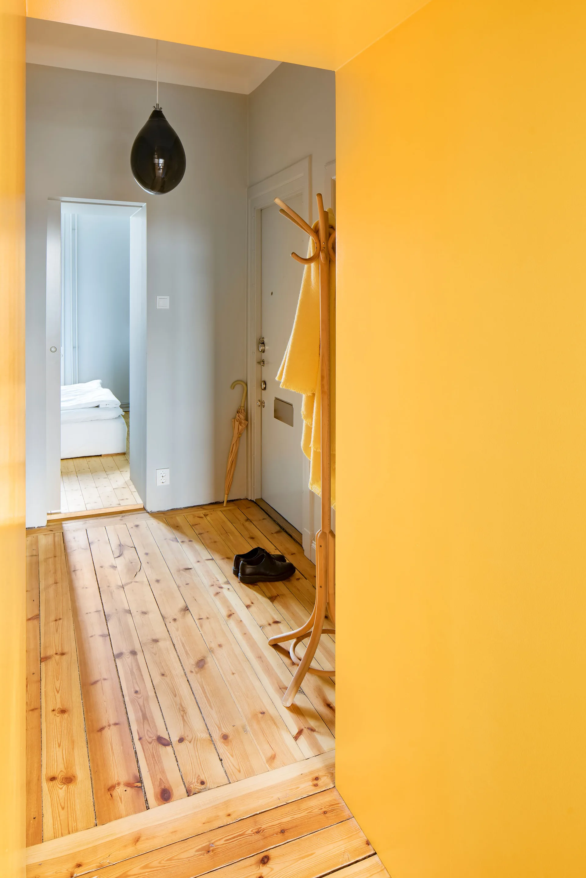 A bright yellow painted wall frames a view into a gray entryway featuring light wood floors, a wooden coat rack, and a dark pendant light.