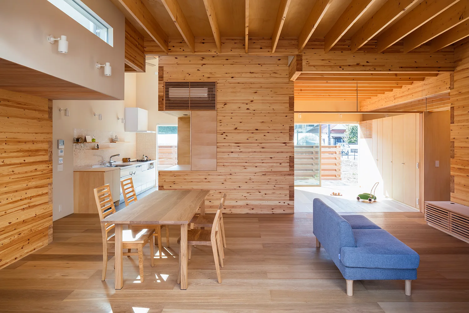 An open-plan wooden interior featuring a dining table, a blue sofa, and exposed ceiling joists.