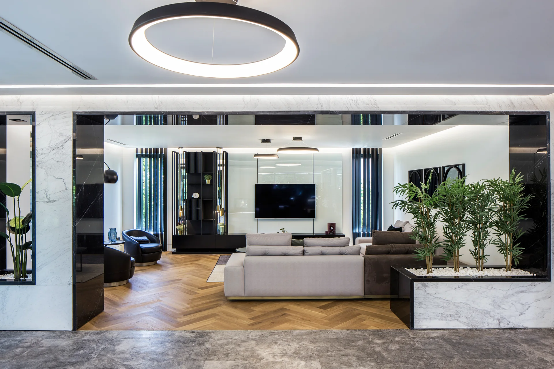 A contemporary living room with chevron wood floors, a grey sectional sofa, and white marble wall paneling framed by a dark mirrored opening.
