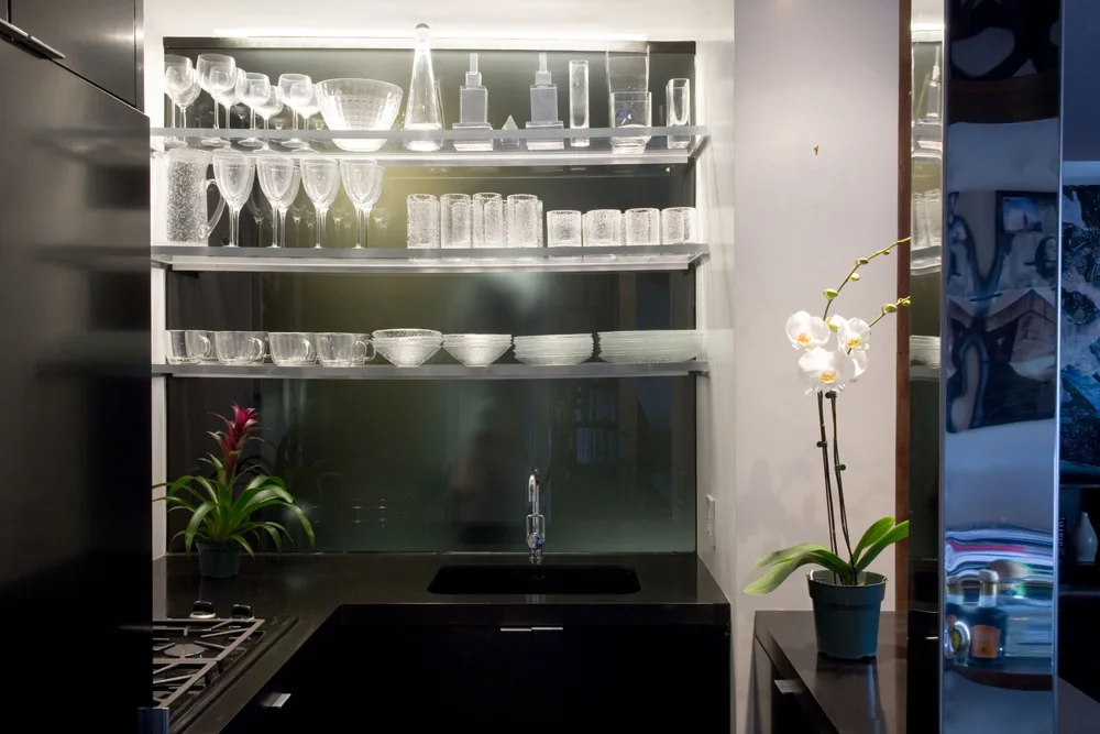 A modern black wet bar featuring illuminated glass shelving stacked with glassware, a dark integrated sink, and a small potted orchid.