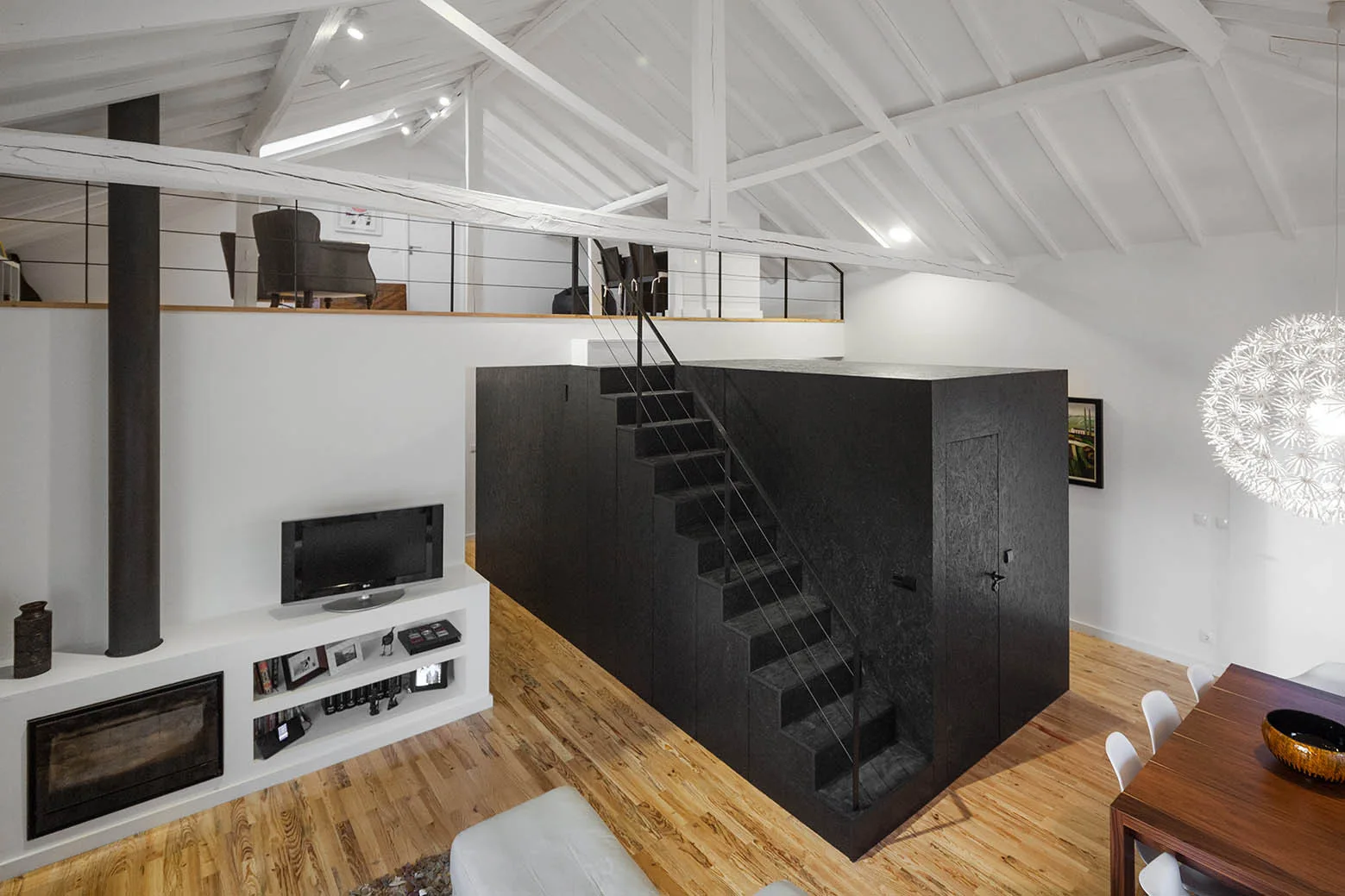 A modern loft interior featuring a matte black staircase block, white vaulted ceilings with exposed beams, and light wood plank flooring.