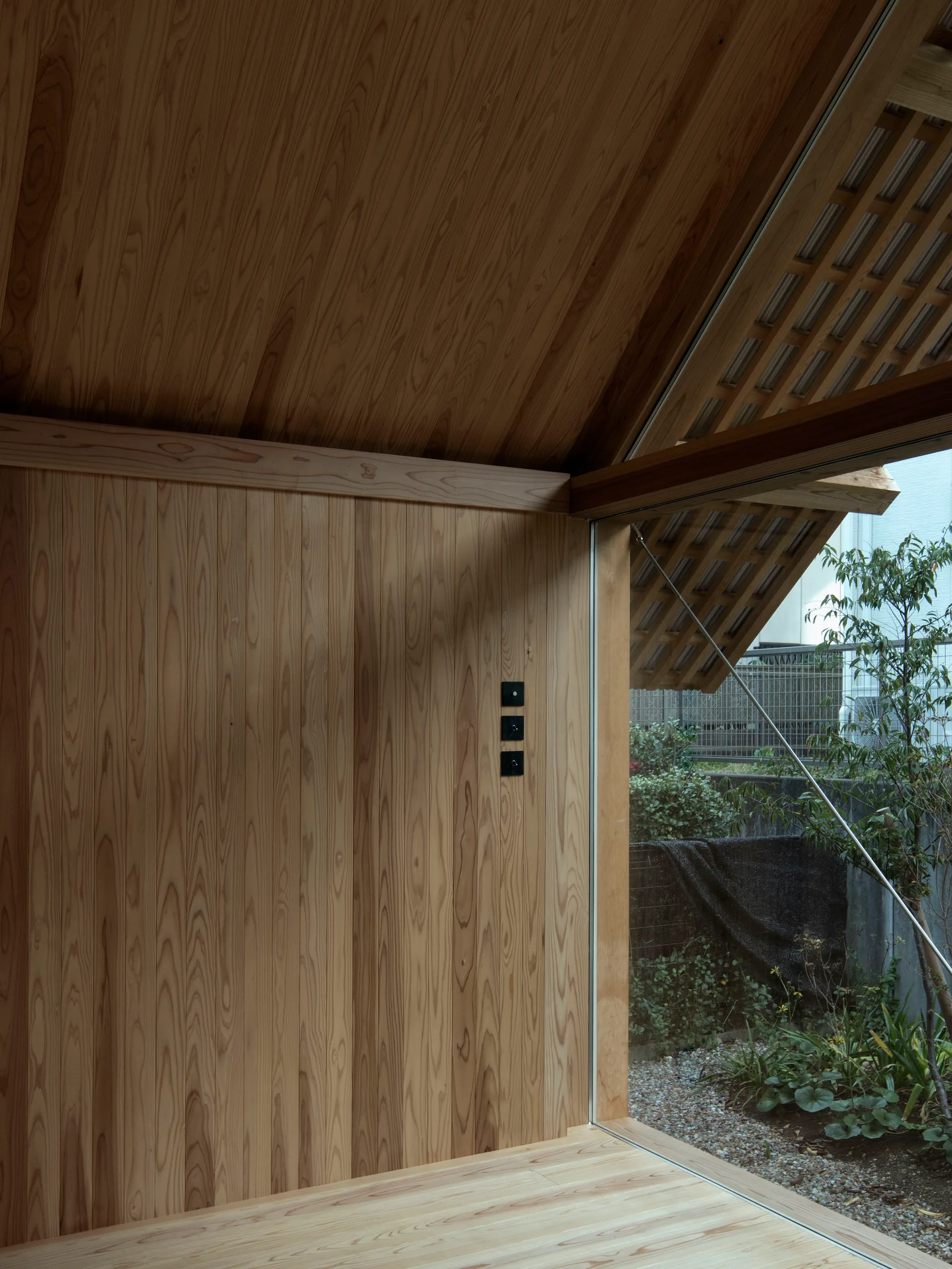 A minimalist room with vertical wood paneling on the walls and angled ceiling features a large glass window opening to a gravel garden.