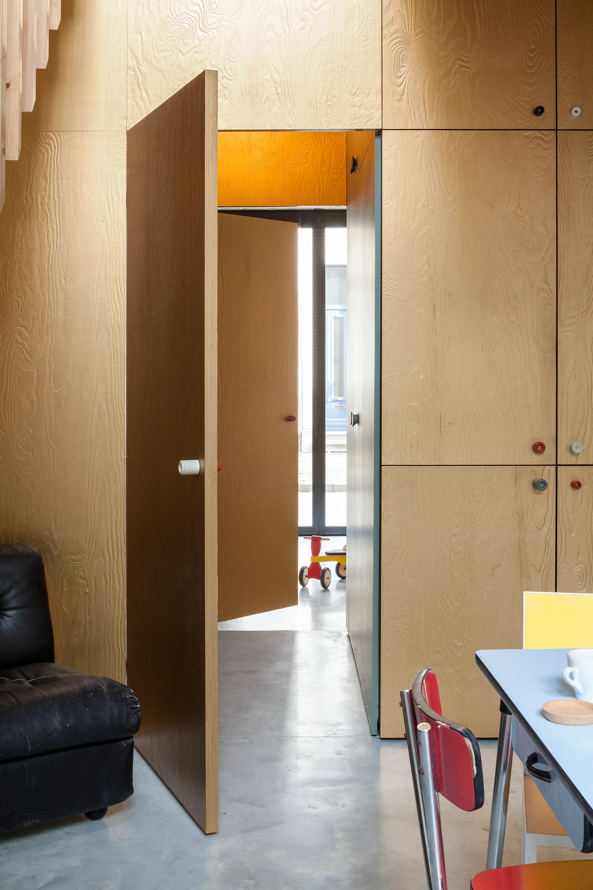 An interior hallway features light tan plywood paneling, open pivoting doors, concrete flooring, and eclectic furniture in the foreground.