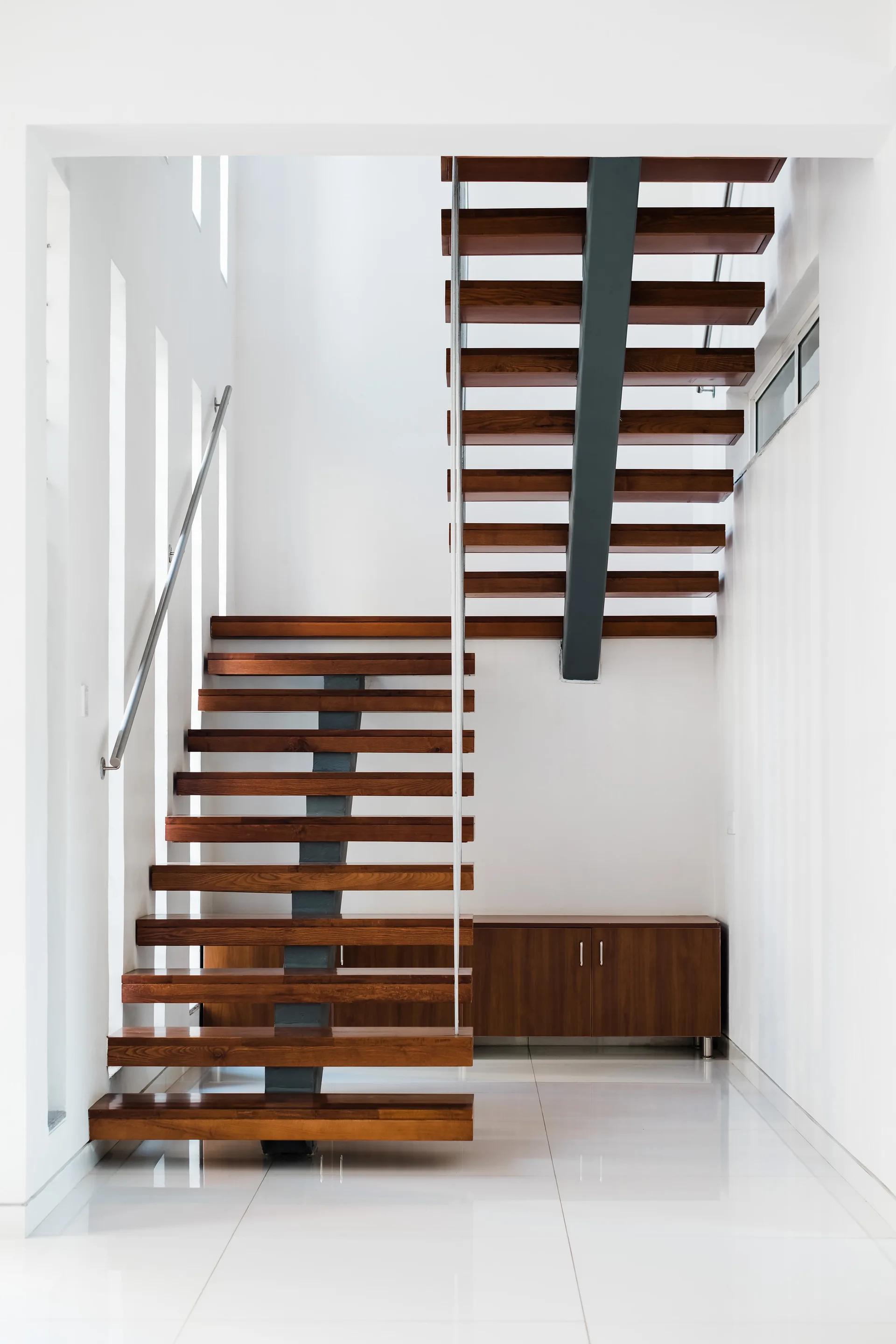 A modern open-riser staircase with walnut treads and a steel spine sits against white walls on a polished tile floor.