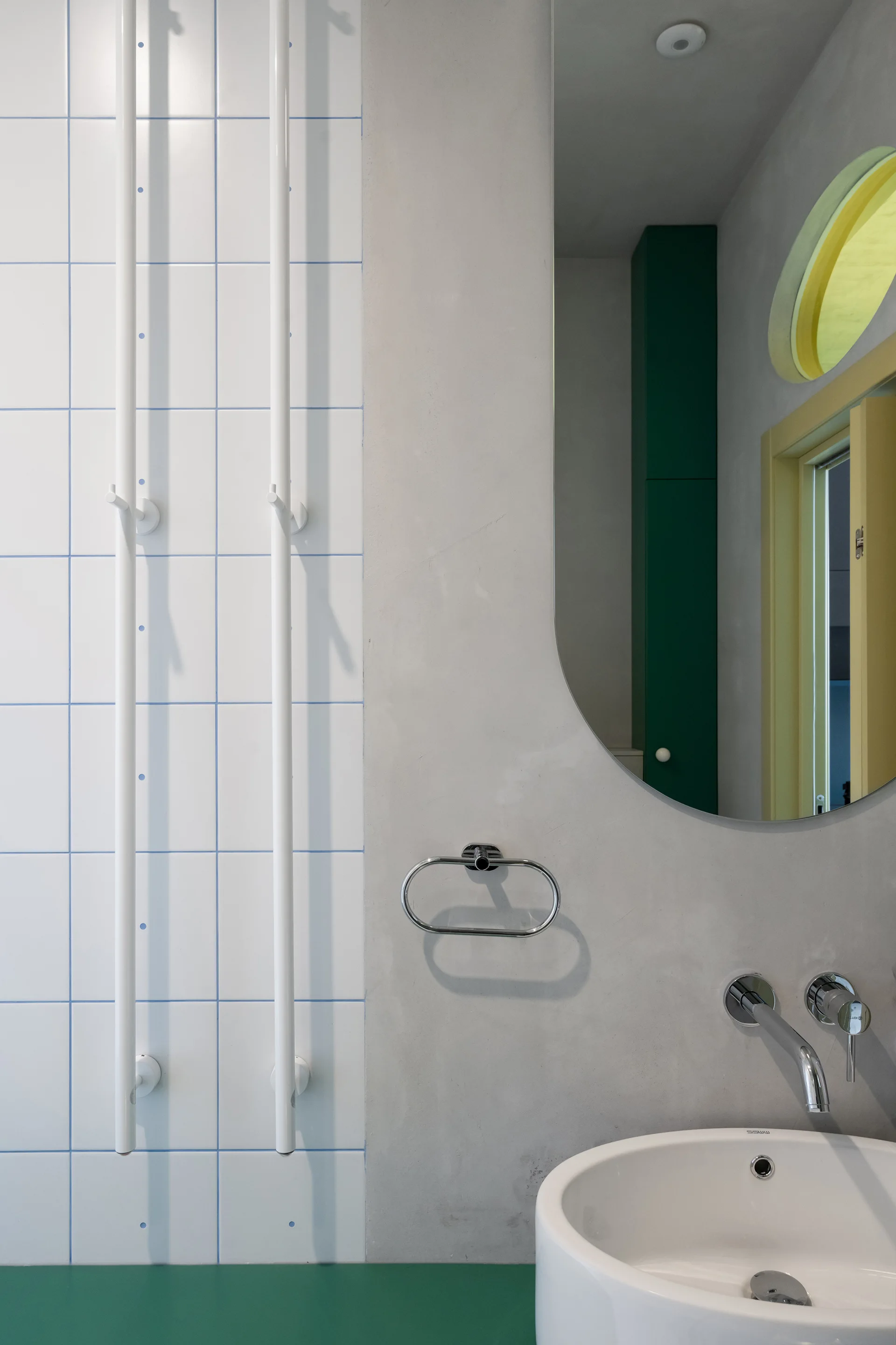 A modern bathroom vanity features a white ceramic sink, wall-mounted faucet, and an oval mirror set against white square tiles with blue grout.