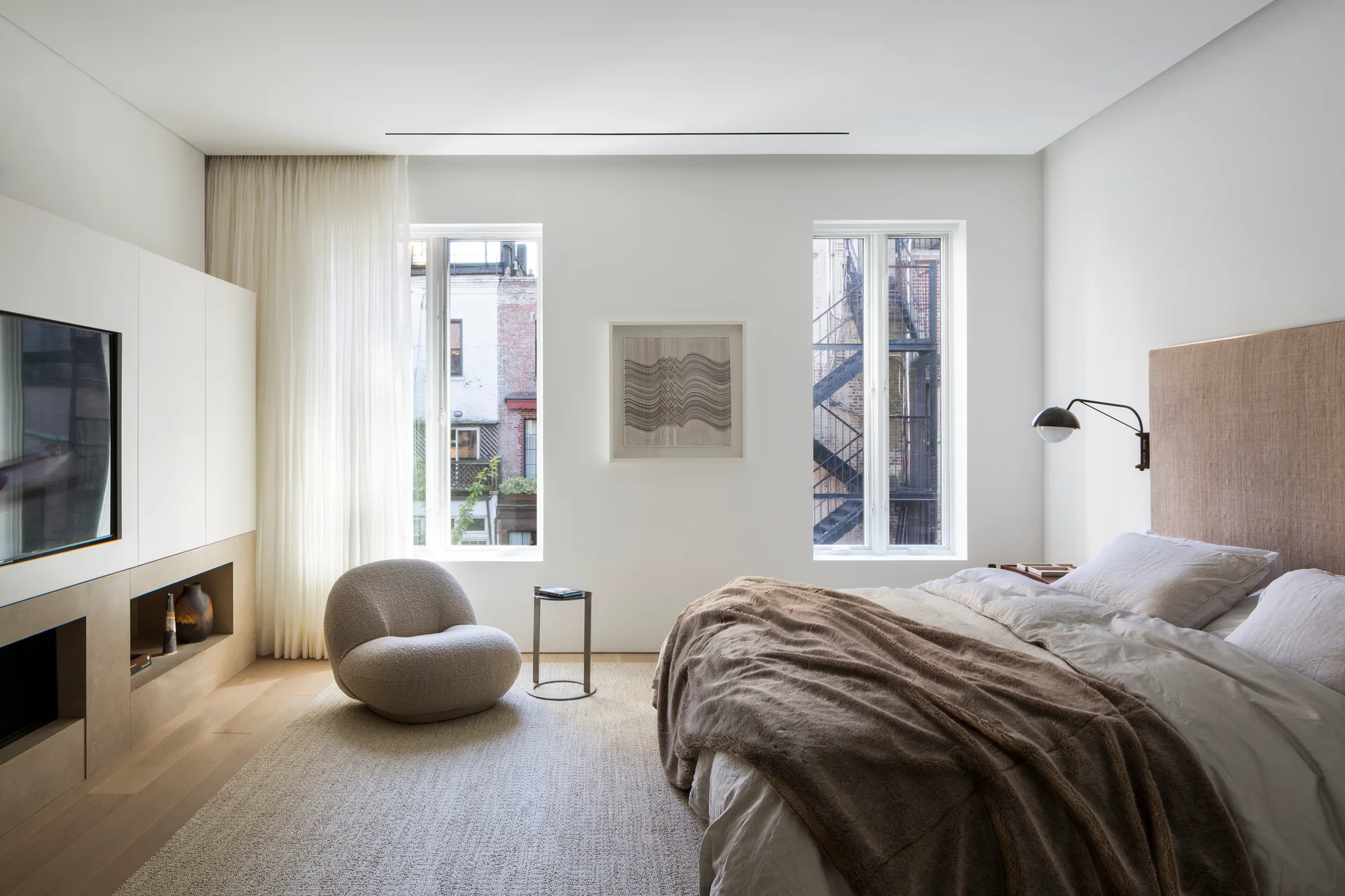 A minimalist bedroom featuring a bed with a woven beige headboard, a curved bouclé armchair near two tall windows, and white built-in shelving.
