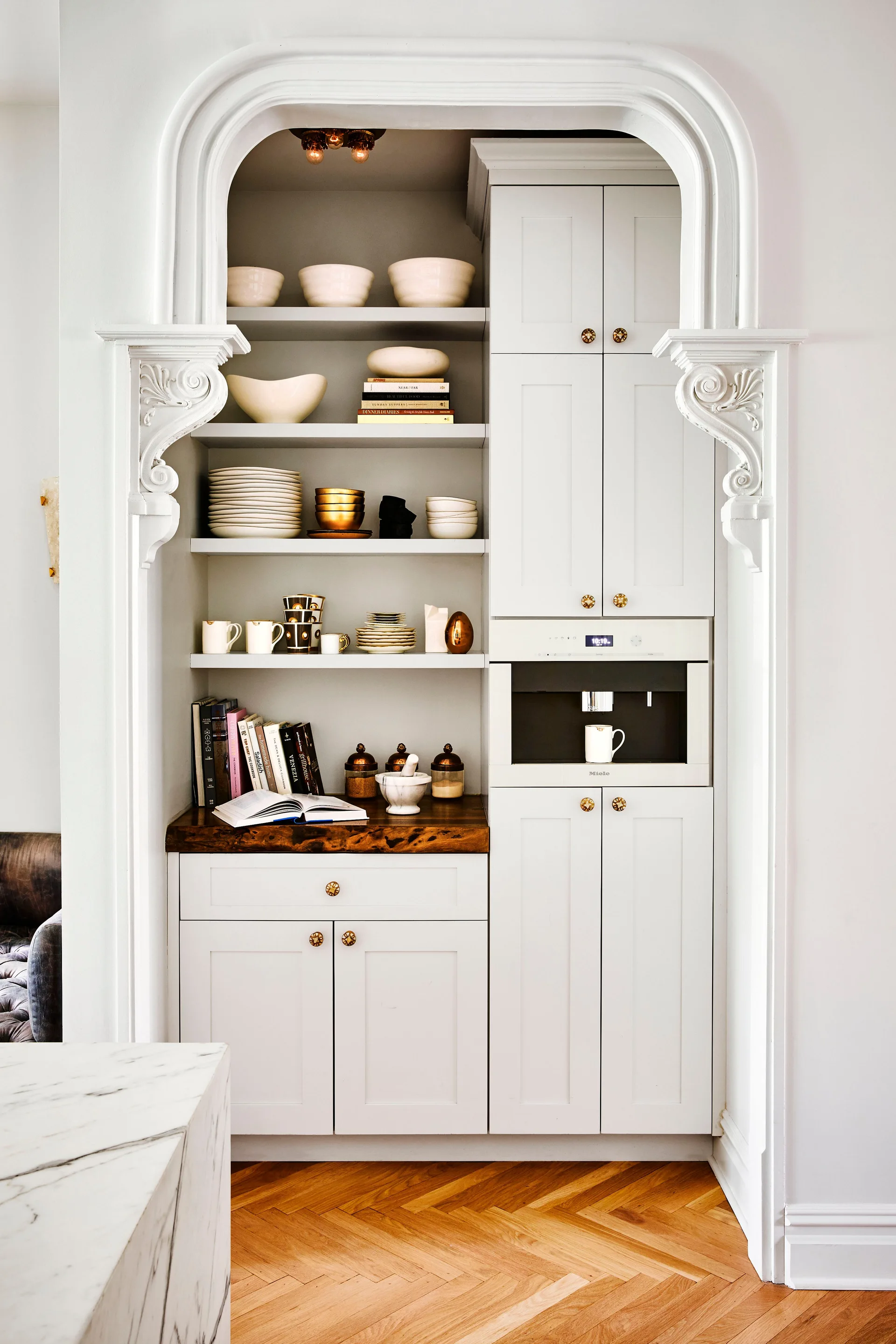 White shaker cabinets and open shelving are built into an ornate arched kitchen nook with herringbone wood flooring.