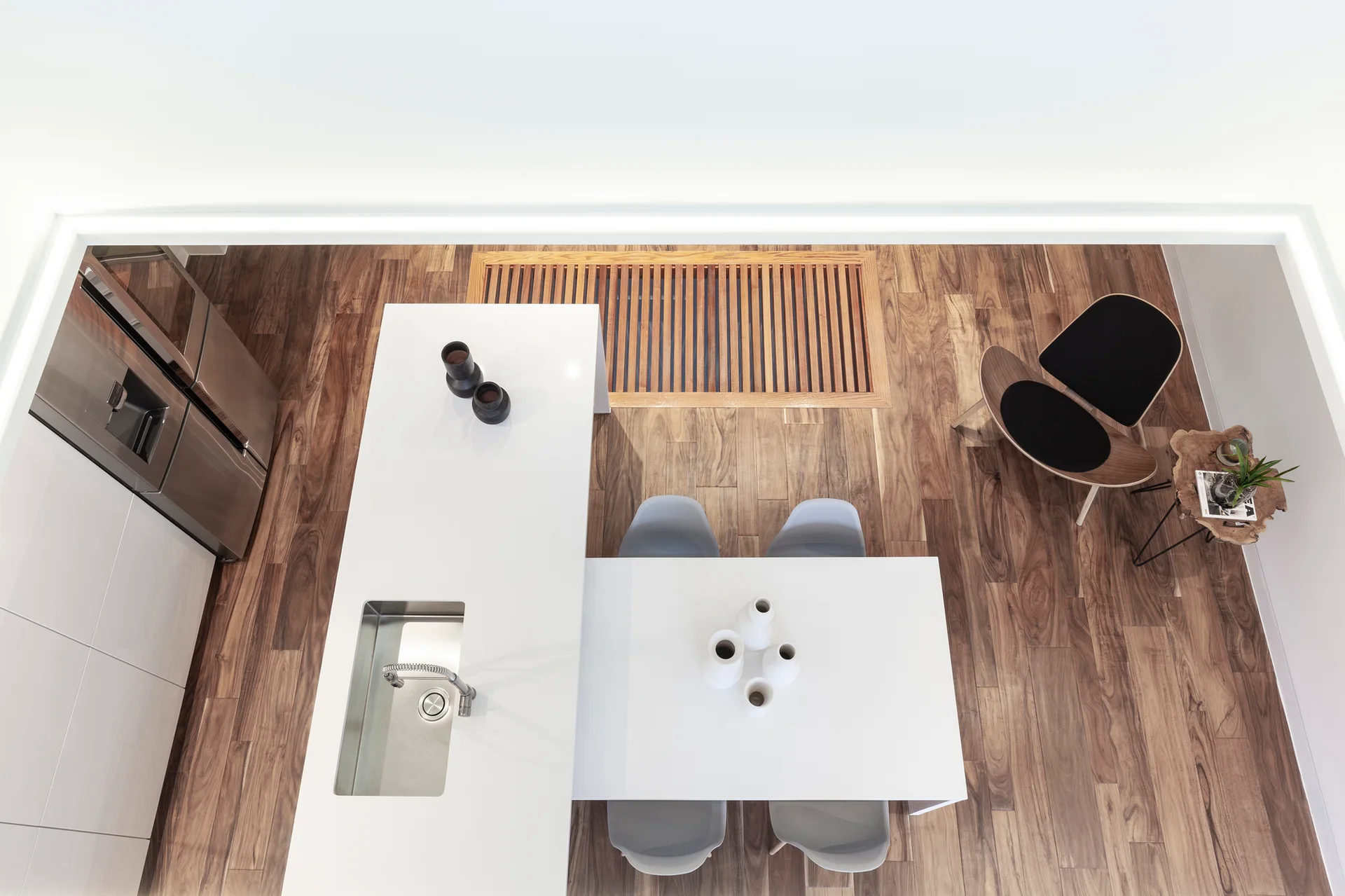Overhead view of a minimalist kitchen featuring a white island with sink, an attached white dining table, grey chairs, and rich hardwood flooring.
