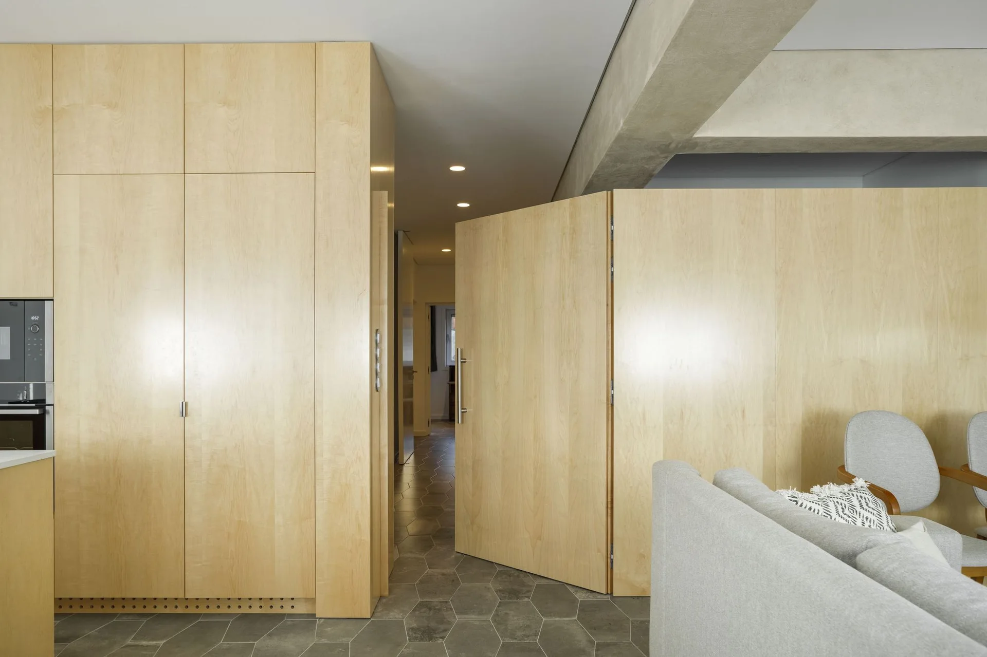 A modern interior features light wood paneled cabinets and a matching pivot door alongside a grey hexagonal tile floor and a grey fabric sofa.