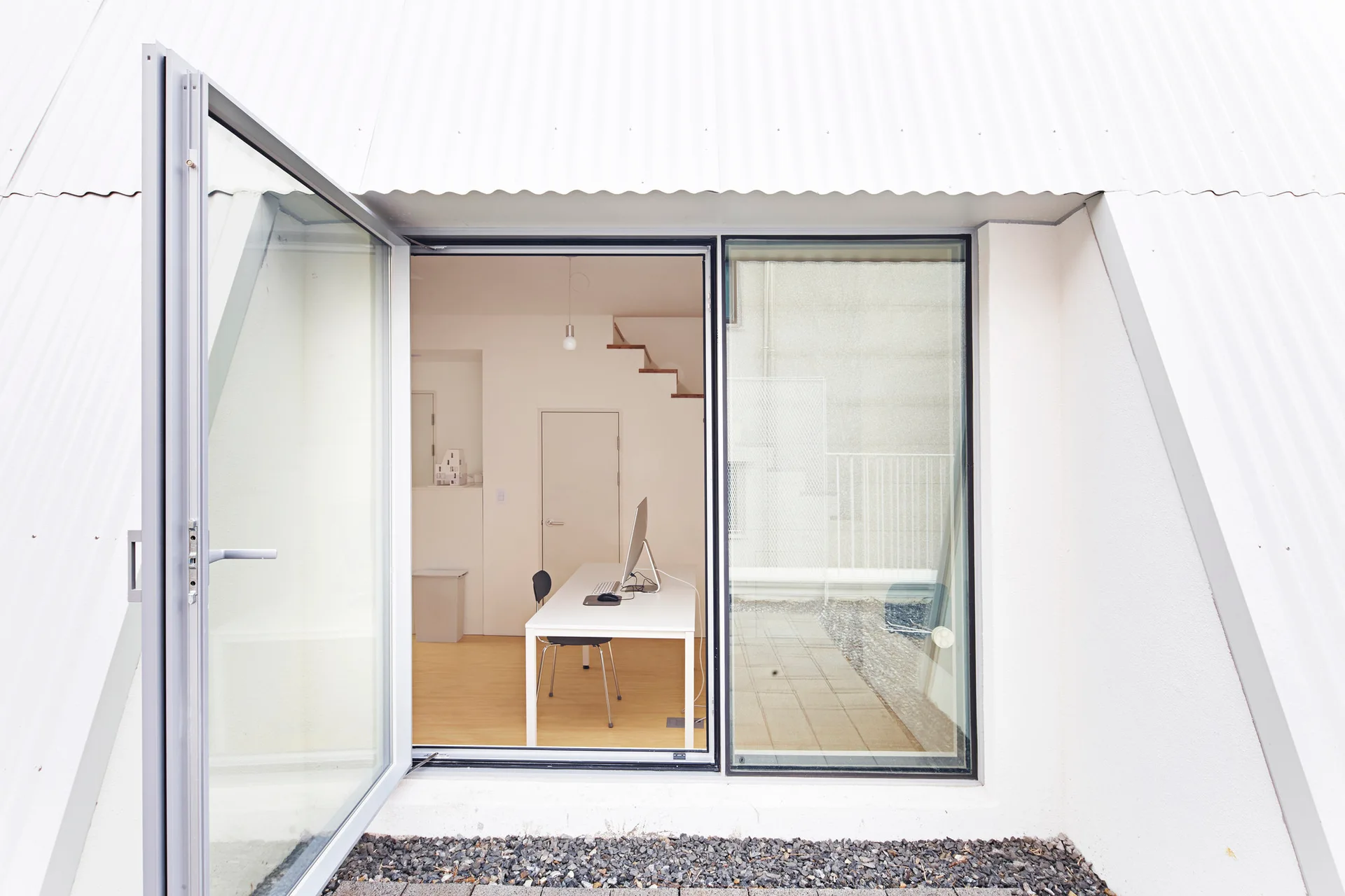 A minimalist home office with a white desk and computer is viewed through a large open glass door.