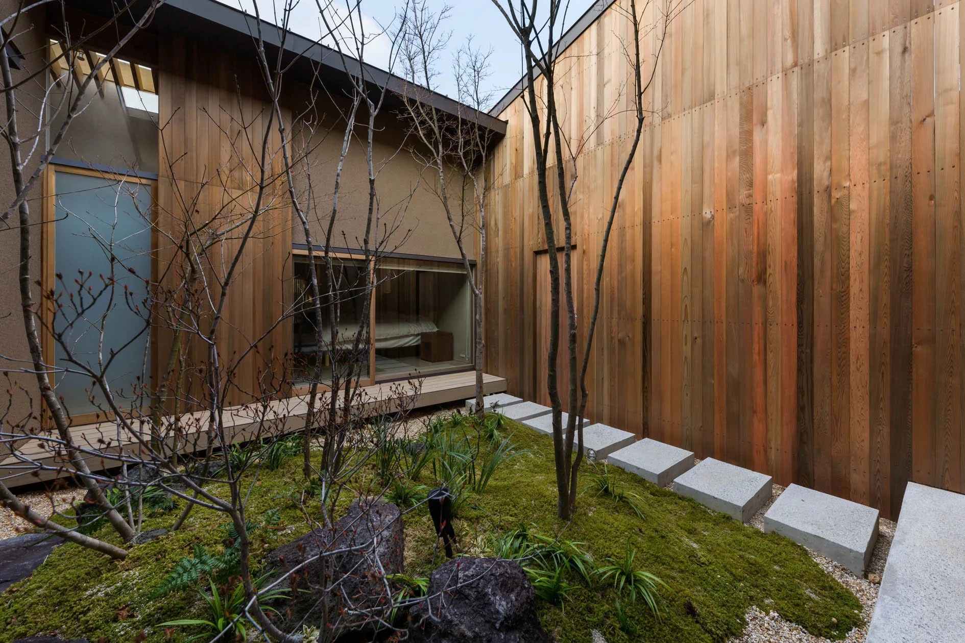 A minimalist courtyard garden features vertical wood siding, mossy ground cover, and a linear path of square concrete stepping stones.