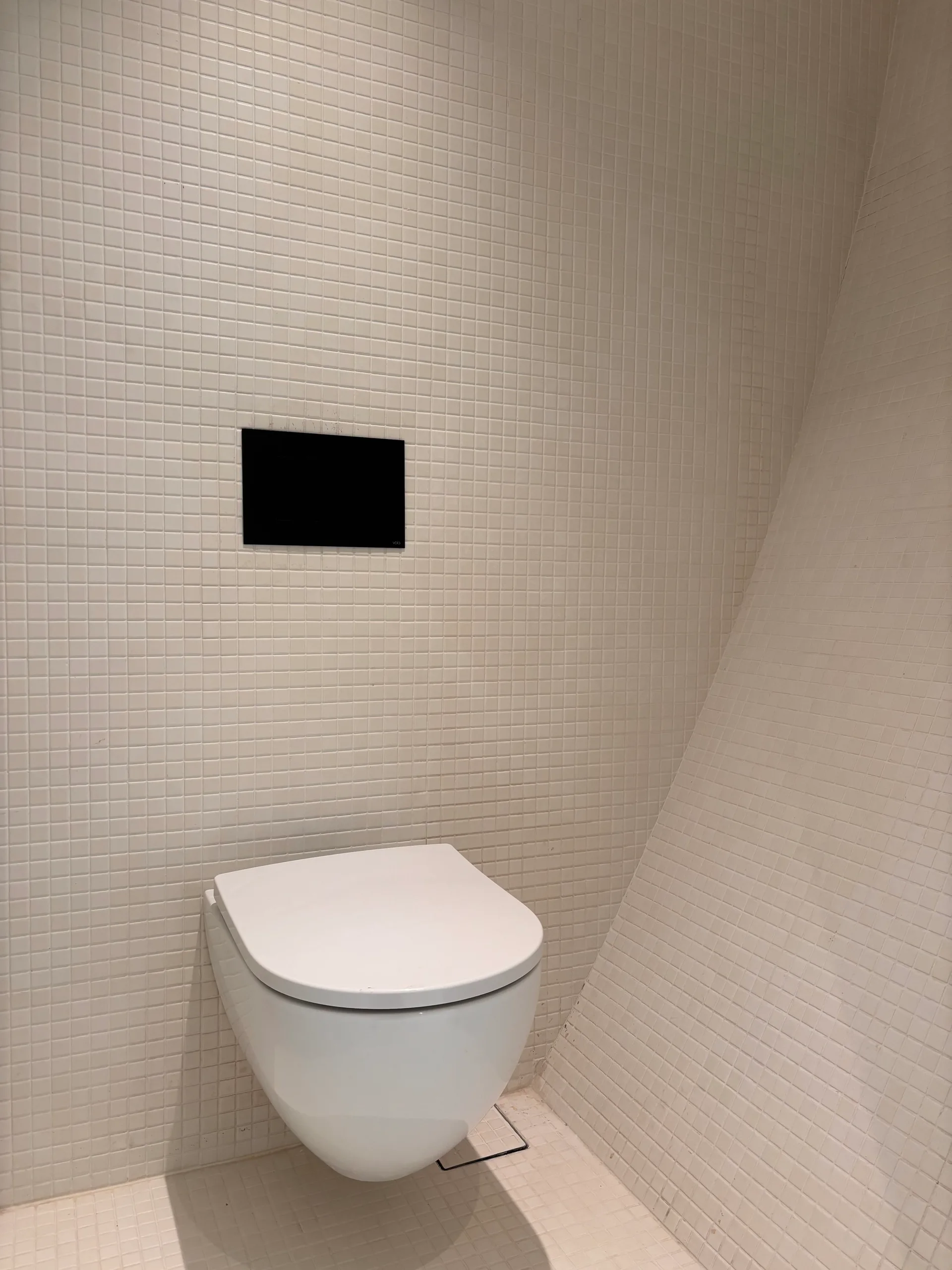 A wall-mounted white ceramic toilet on a wall of small white square mosaic tiles beneath a rectangular black flush plate.