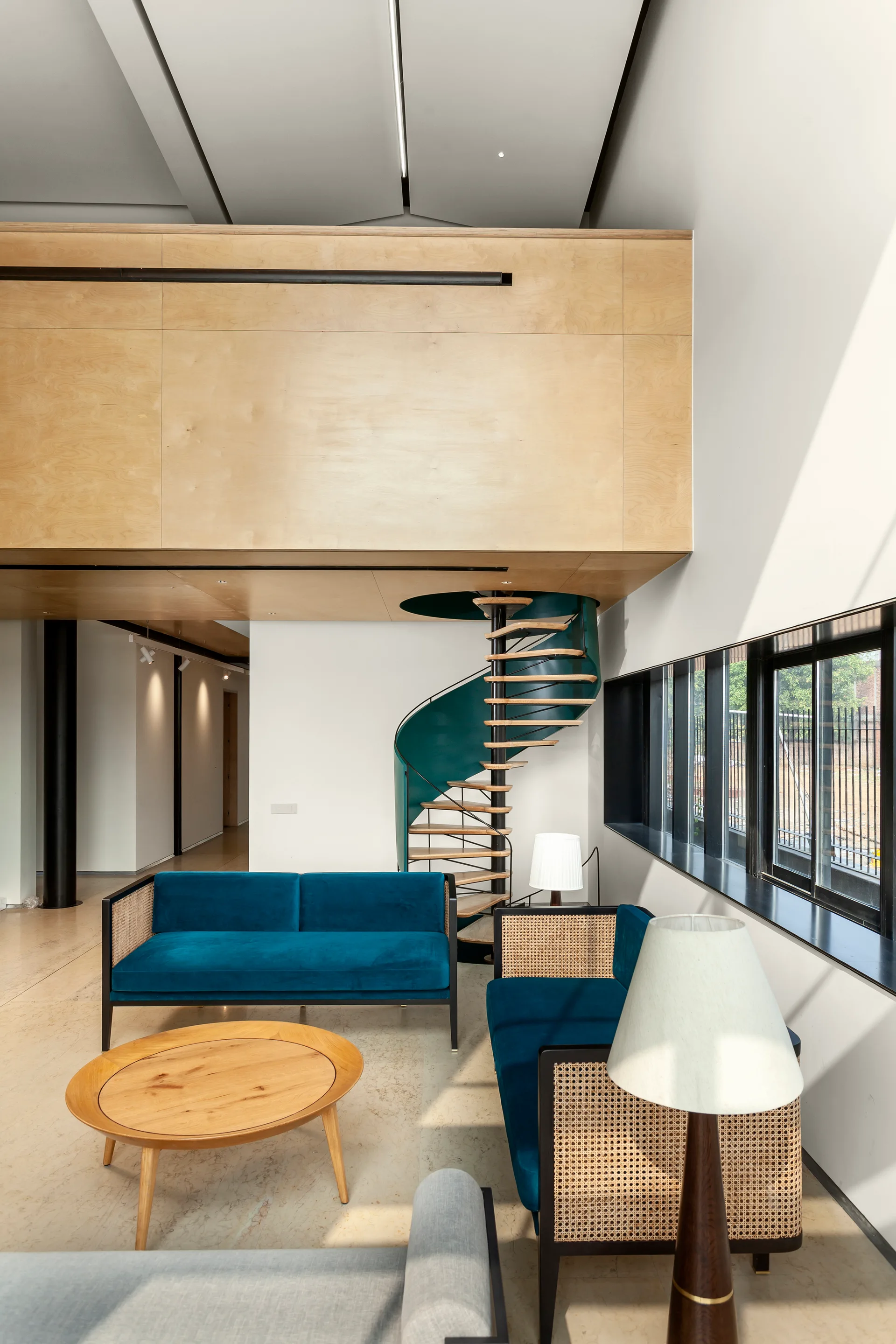 A modern living room features blue velvet sofas with cane siding, a light wood coffee table, and a dark green spiral staircase.