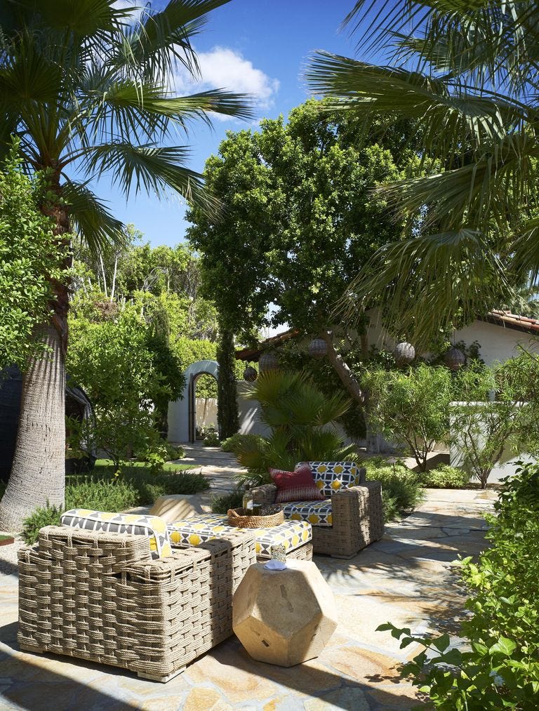 Woven wicker armchairs with yellow geometric cushions sit on an irregular flagstone patio surrounded by lush green palm trees.