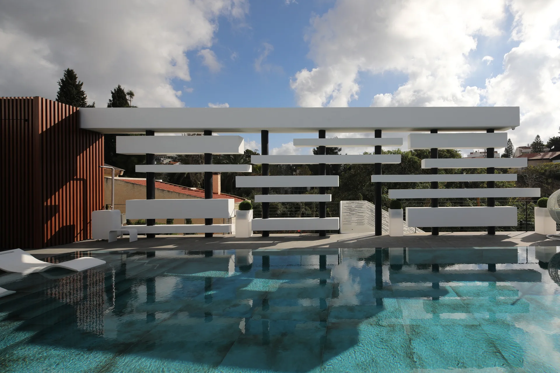 A modern swimming pool is bordered by a white geometric sculptural screen and a brown slatted accent wall under a cloudy sky.